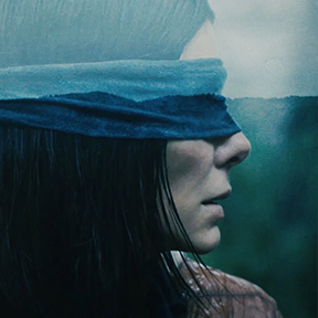A person with long dark hair wearing a blindfold, viewed from the side against a blurred natural background.