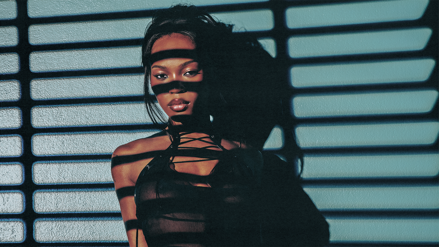 Quenlin Blackwell stands against a wall with shadows of blinds casting lines across her face and body, wearing a black top.