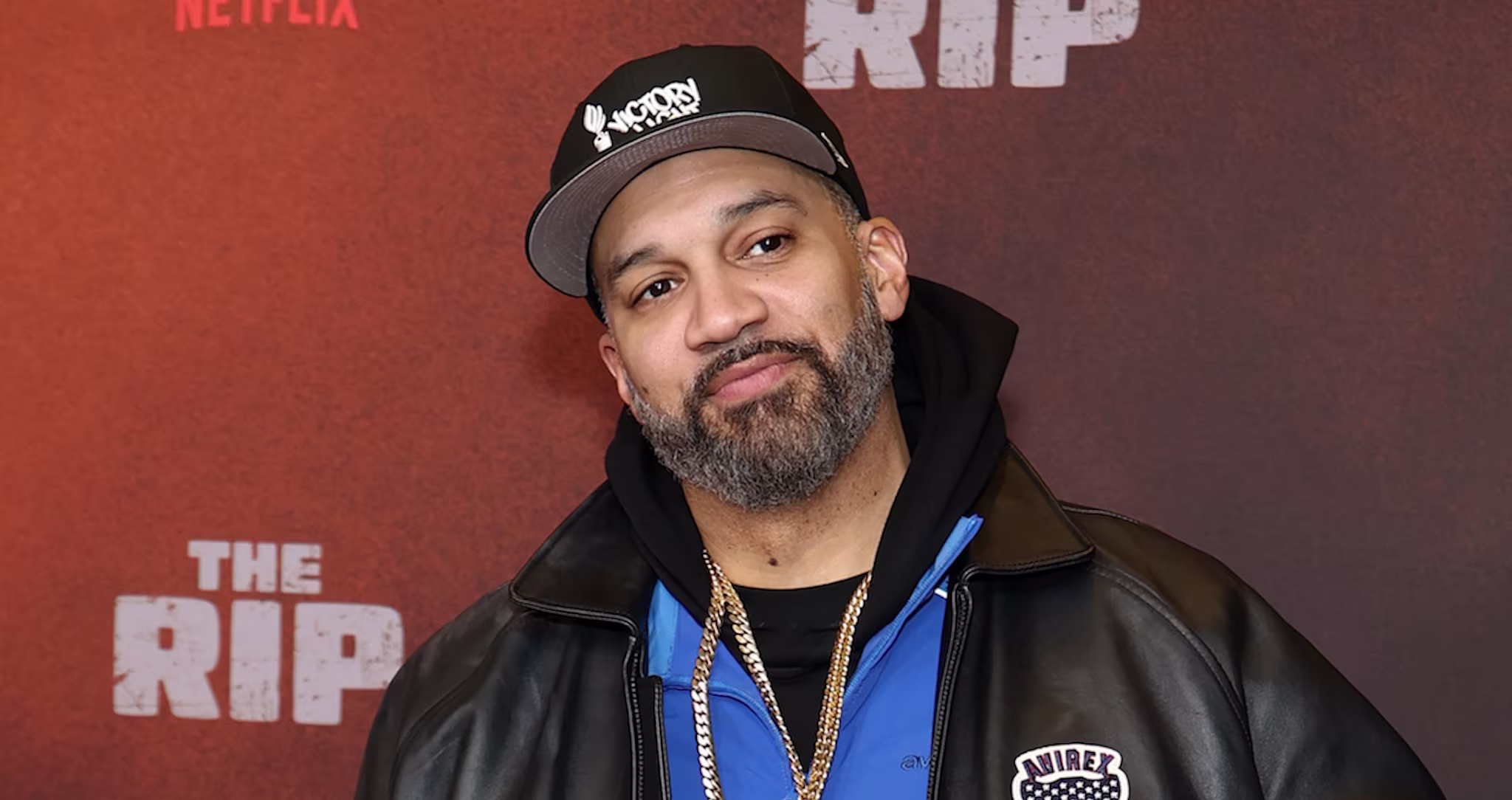 The Kid Mero with a beard wearing a cap and leather jacket stands in front of a backdrop.
