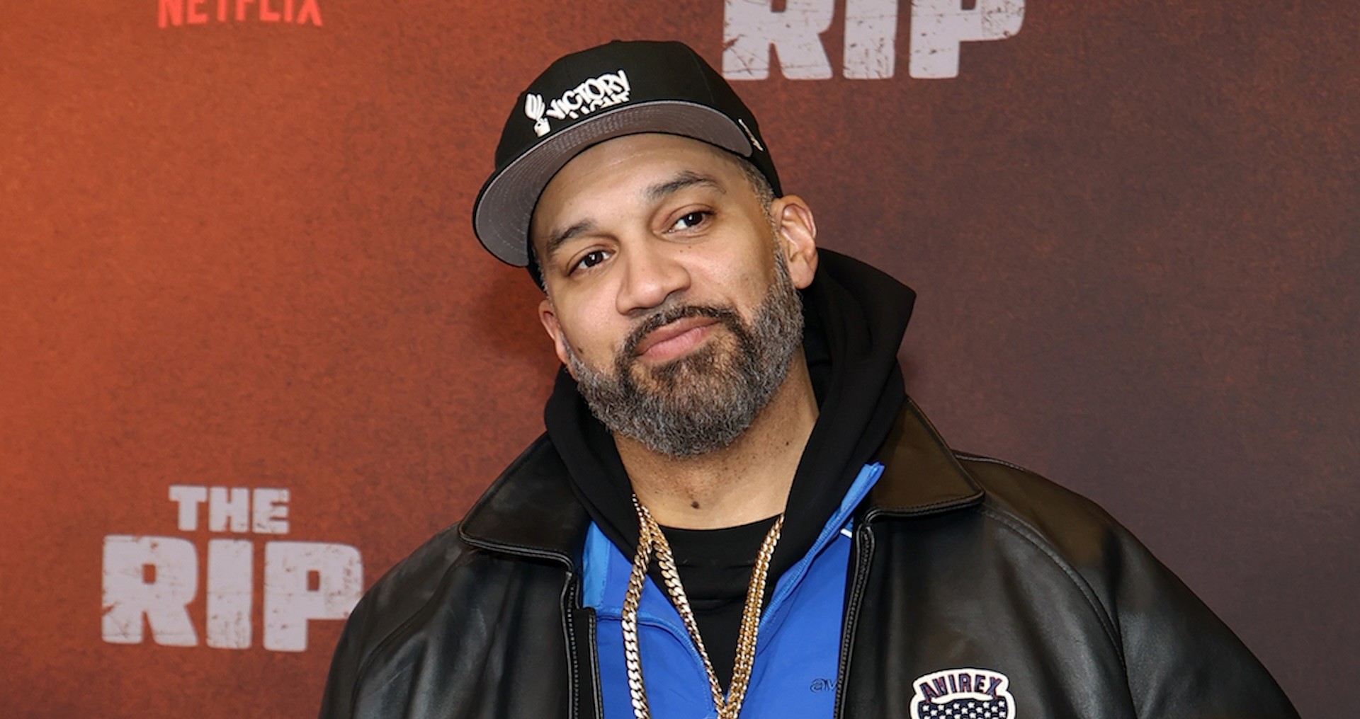 The Kid Mero with a beard wearing a cap and leather jacket stands in front of a backdrop.