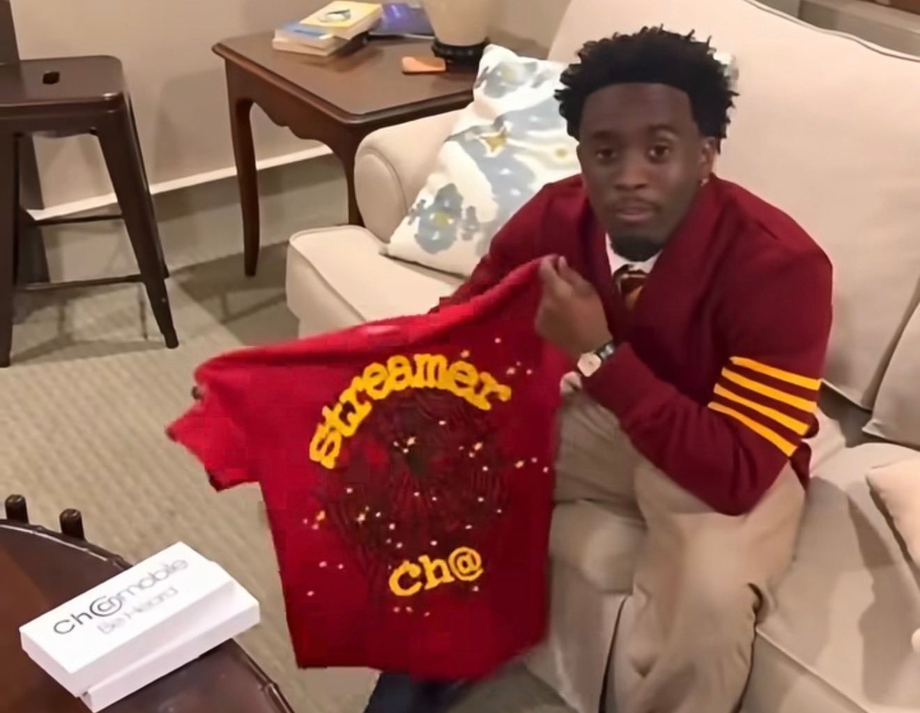 A person sitting on a sofa holding a red shirt with "streamer ch@" written on it. A table with a box and a lamp are nearby.