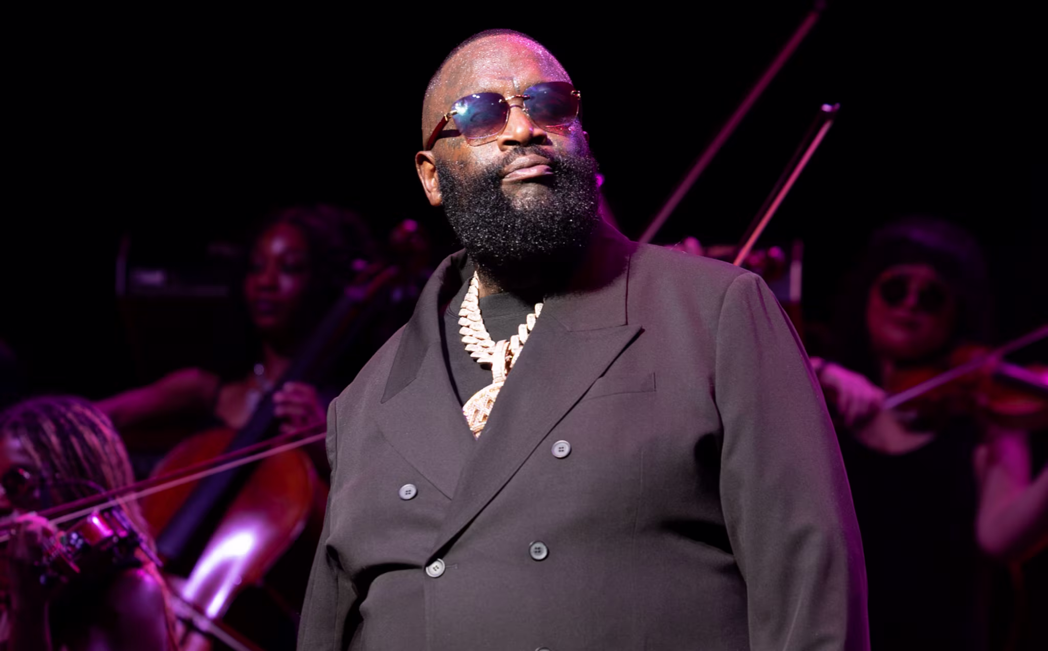 Rick Ross in a black suit and sunglasses performs on stage with musicians playing string instruments in the background.