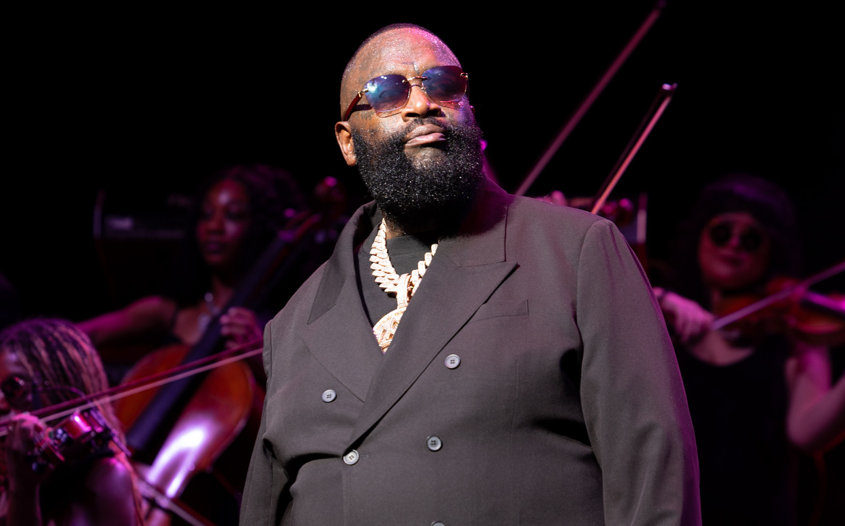 Rick Ross in a black suit and sunglasses performs on stage with musicians playing string instruments in the background.
