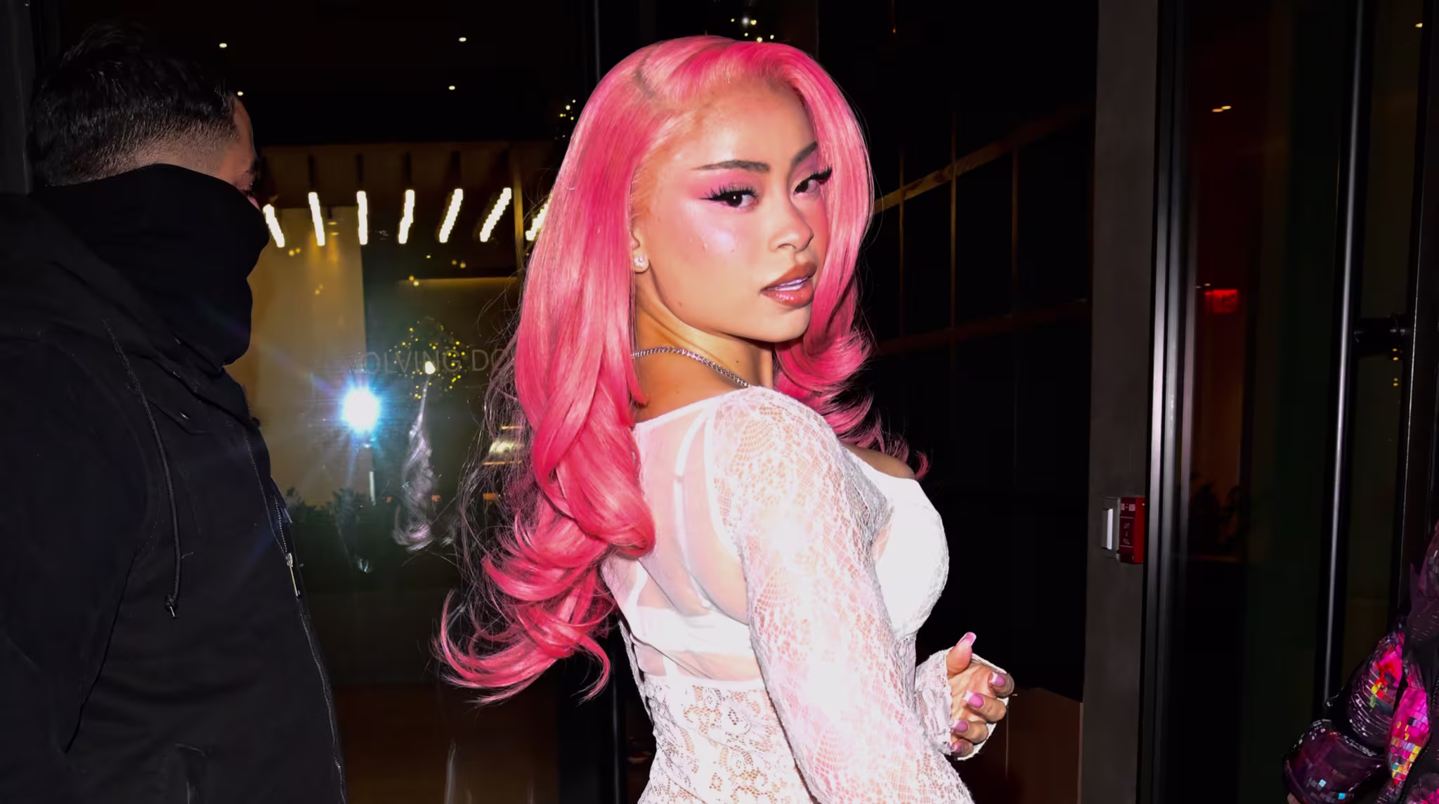 Ice Spice with pink hair in a sheer white lace dress poses for a photo. Two people stand nearby in a dimly lit entrance.