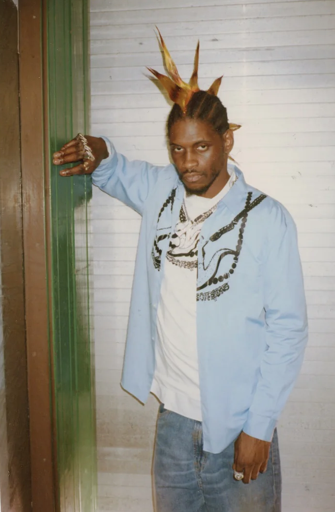 ASAP Nast with unique spiked hairstyle, wearing a light blue shirt over a white t-shirt and jeans, leaning against a wall.