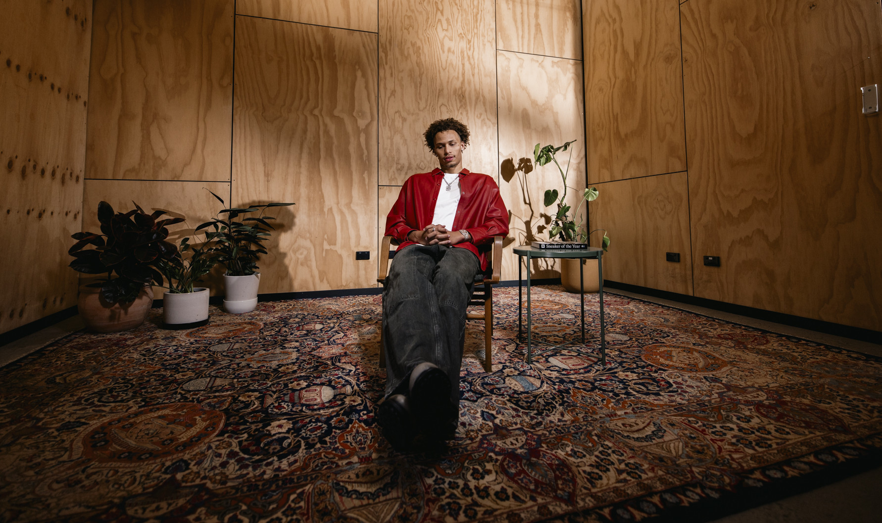 Dyson in a red jacket sits on a chair in a wooden room with a patterned rug, surrounded by plants and a small table.