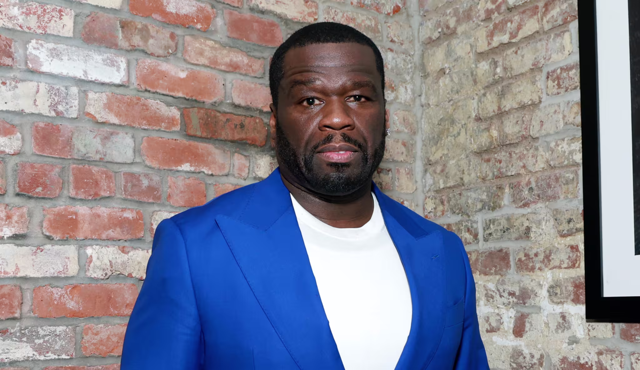 50 Cent in a bright blue suit and white shirt, standing against a brick wall with framed artwork and candles on a shelf.