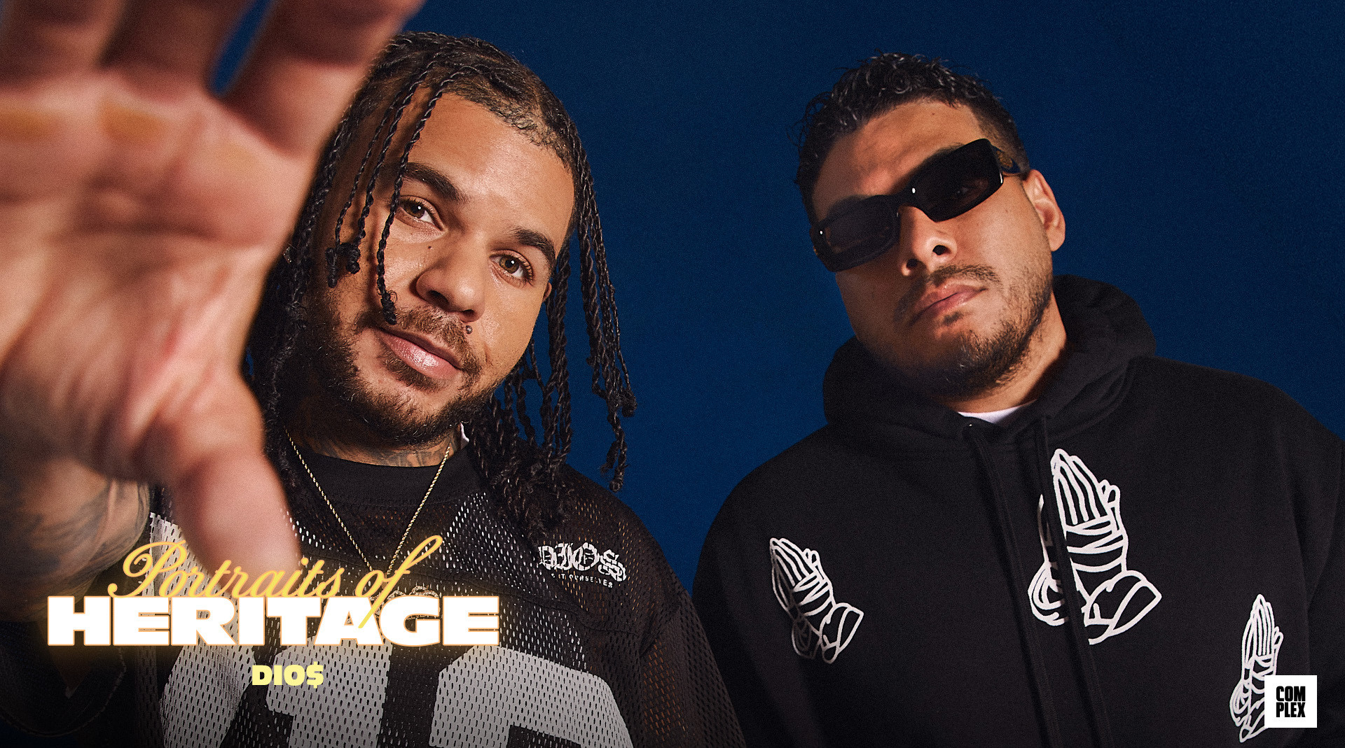 Two men pose against a blue background, one with braids reaching forward, the other wearing sunglasses and a hoodie with praying hands.