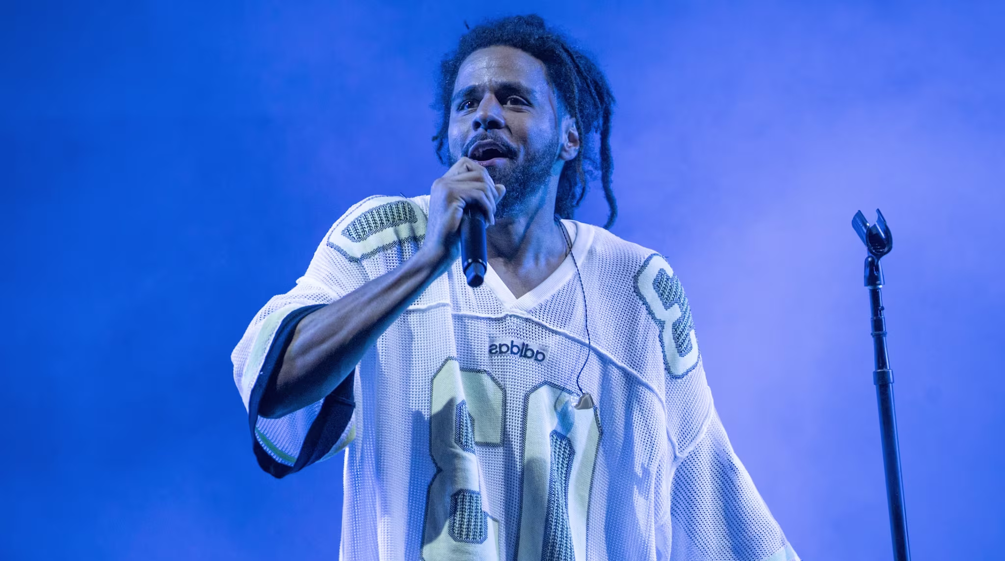 J. Cole performing on stage in a sports jersey, holding a microphone against a blue-lit background.