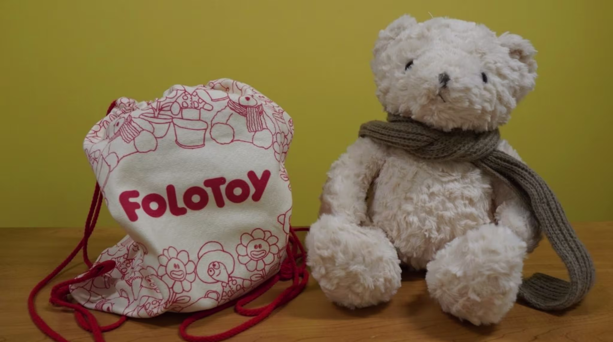 A plush teddy bear with a scarf sits beside a drawstring bag labeled "Folotoy" with red illustrations.