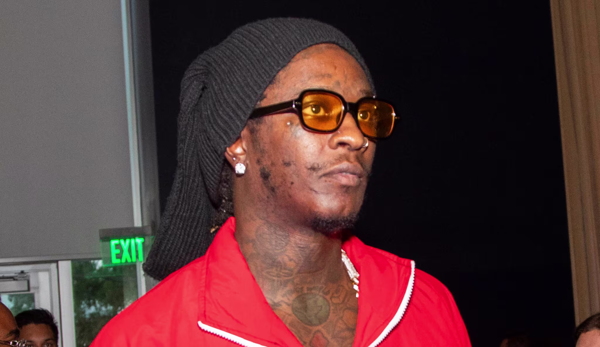Young Thug in a red tracksuit and sunglasses, with a black head covering, stands in a crowd.