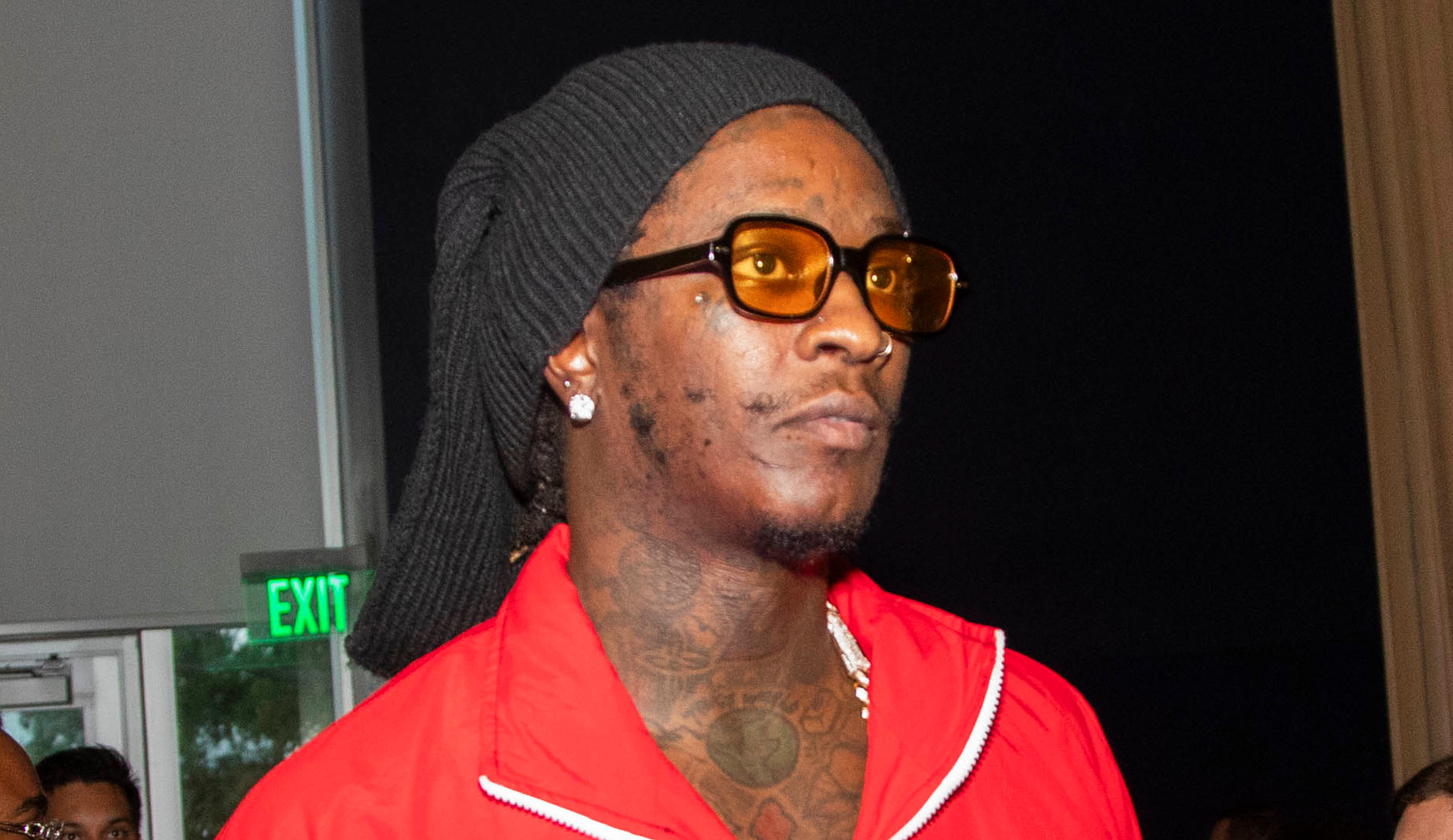 Young Thug in a red tracksuit with a black headscarf and yellow sunglasses at an event, surrounded by people.