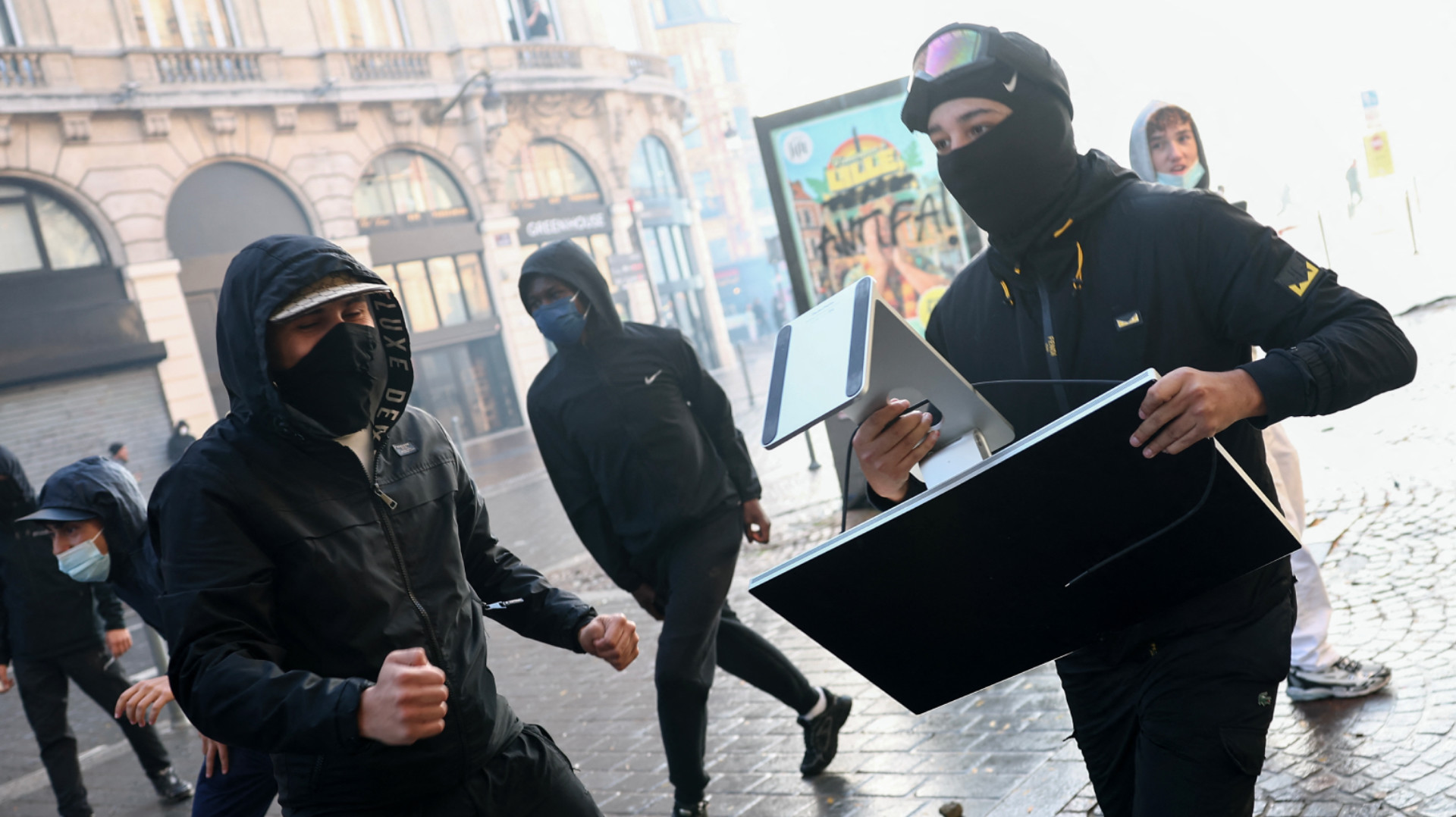 A group of masked individuals in dark clothing, one carrying a computer monitor, walk on a city street with historic buildings in the background.