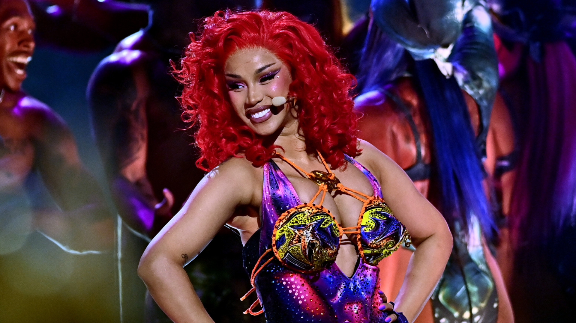 Cardi B performing on stage with vibrant red hair and a colorful, futuristic outfit, smiling confidently.