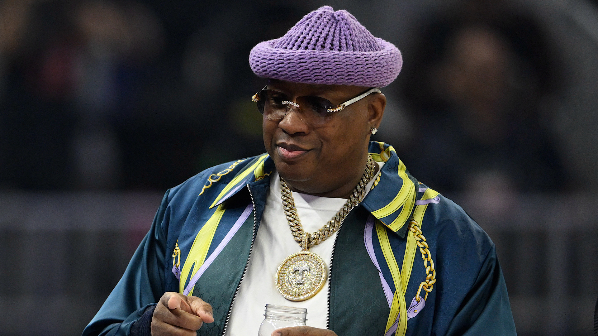 Rapper E-40 wearing a purple knit hat, glasses, and gold chains, holding a drink at an event.
