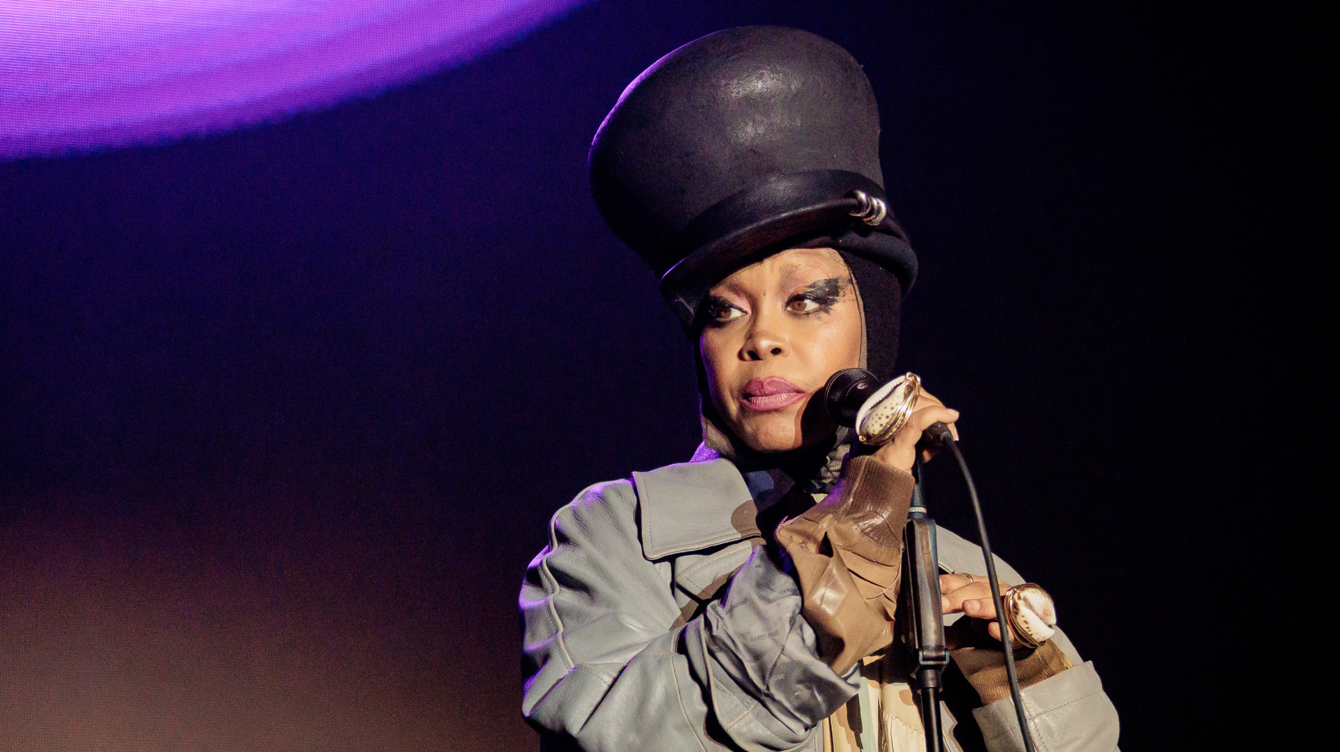 Singer Erykah Badu performing on stage, wearing a large black hat and holding a microphone.