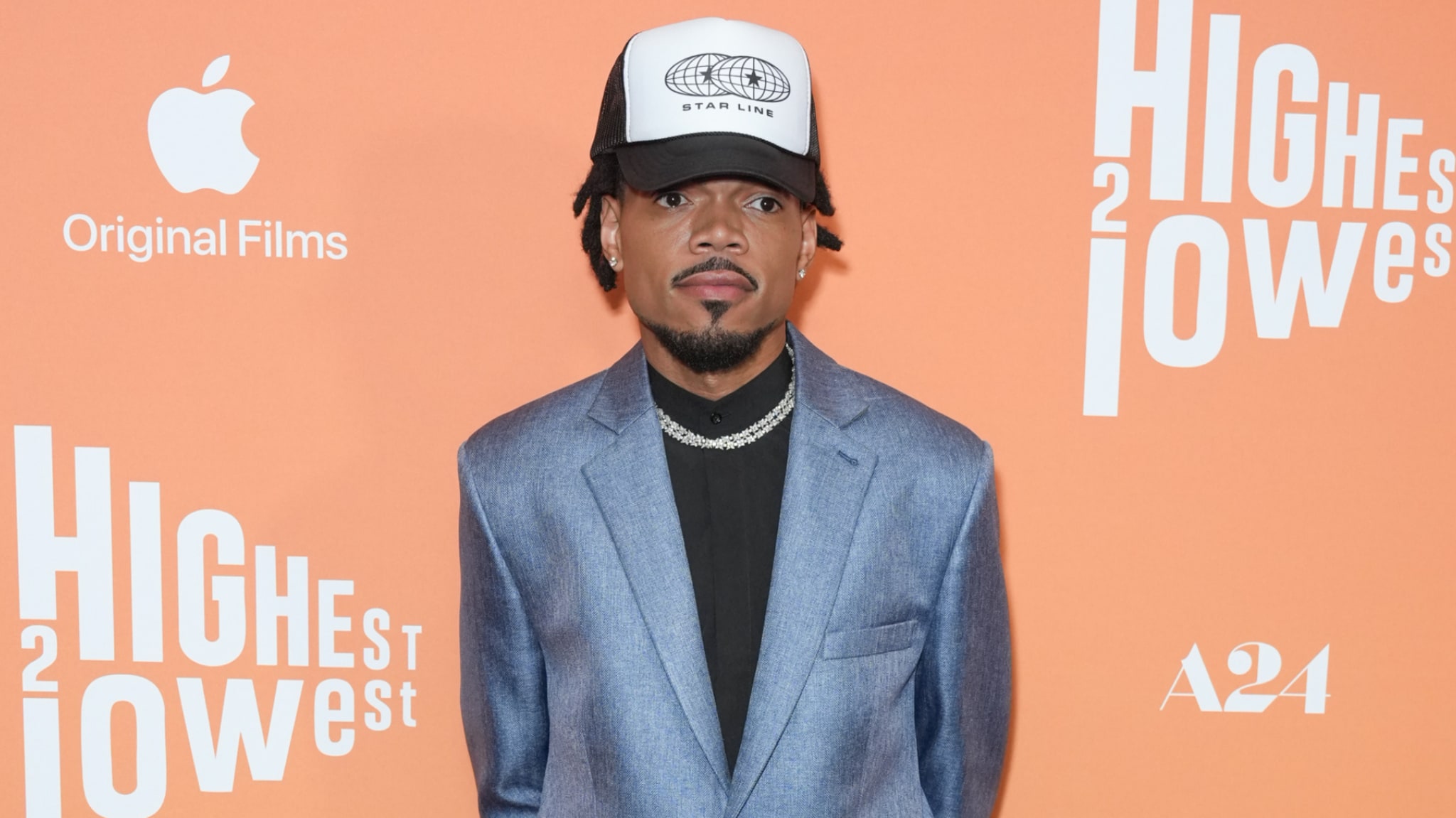Chance the Rapper in a blue suit and cap at an event with Apple and A24 logos on an orange backdrop.