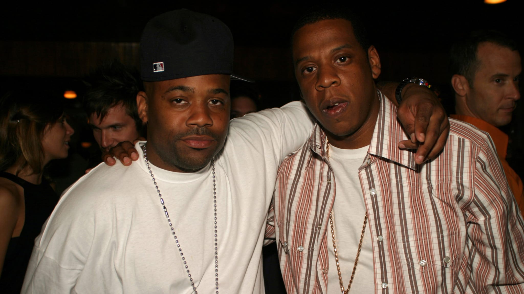Damon Dash and Jay-Z pose together at an event. Damon wears a white shirt and cap, while Jay-Z sports a striped shirt and chain.