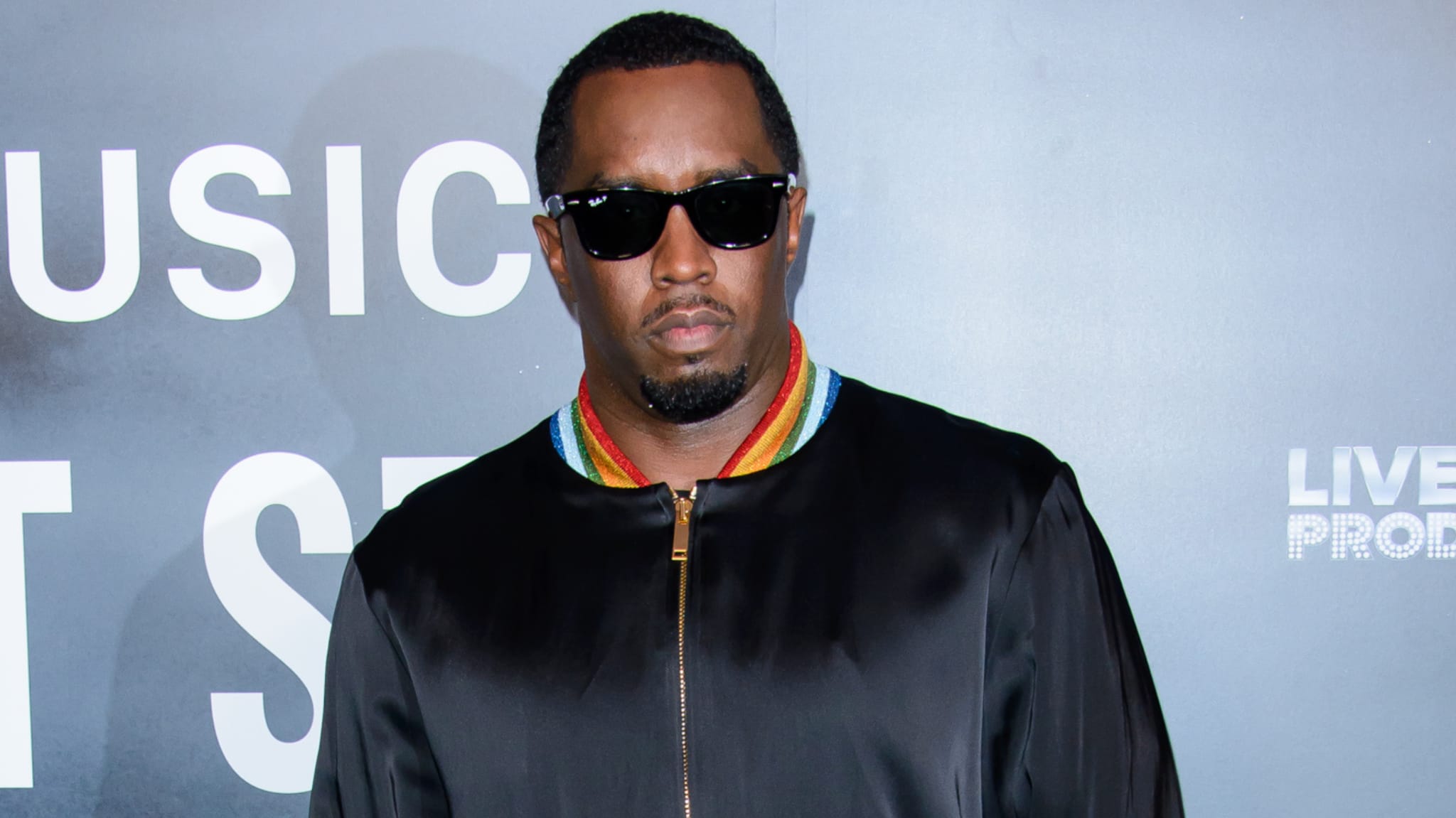 Sean "Diddy" Combs wearing sunglasses and a black jacket with a colorful collar, standing against a backdrop with text.