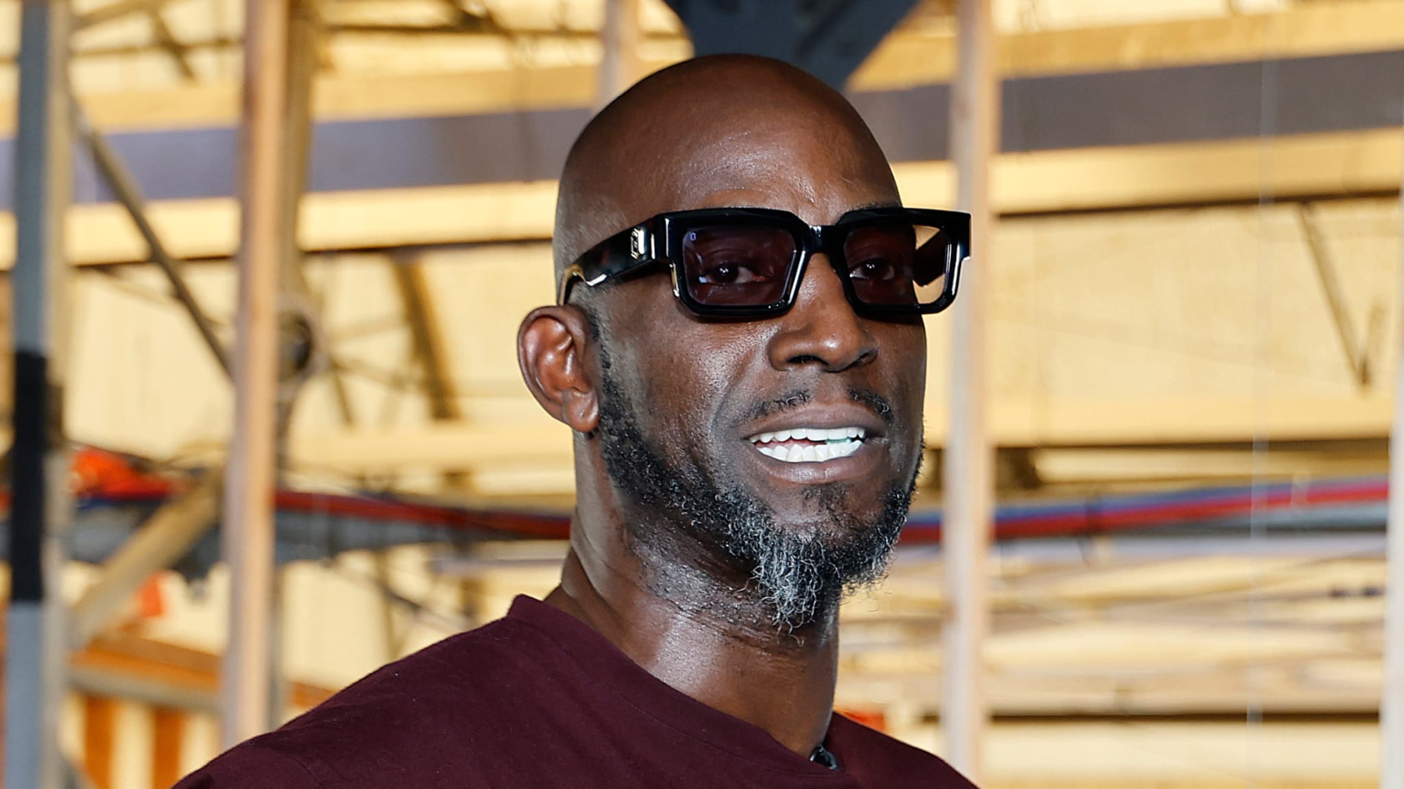Man wearing sunglasses and a maroon shirt, with a beard, standing indoors with industrial background.