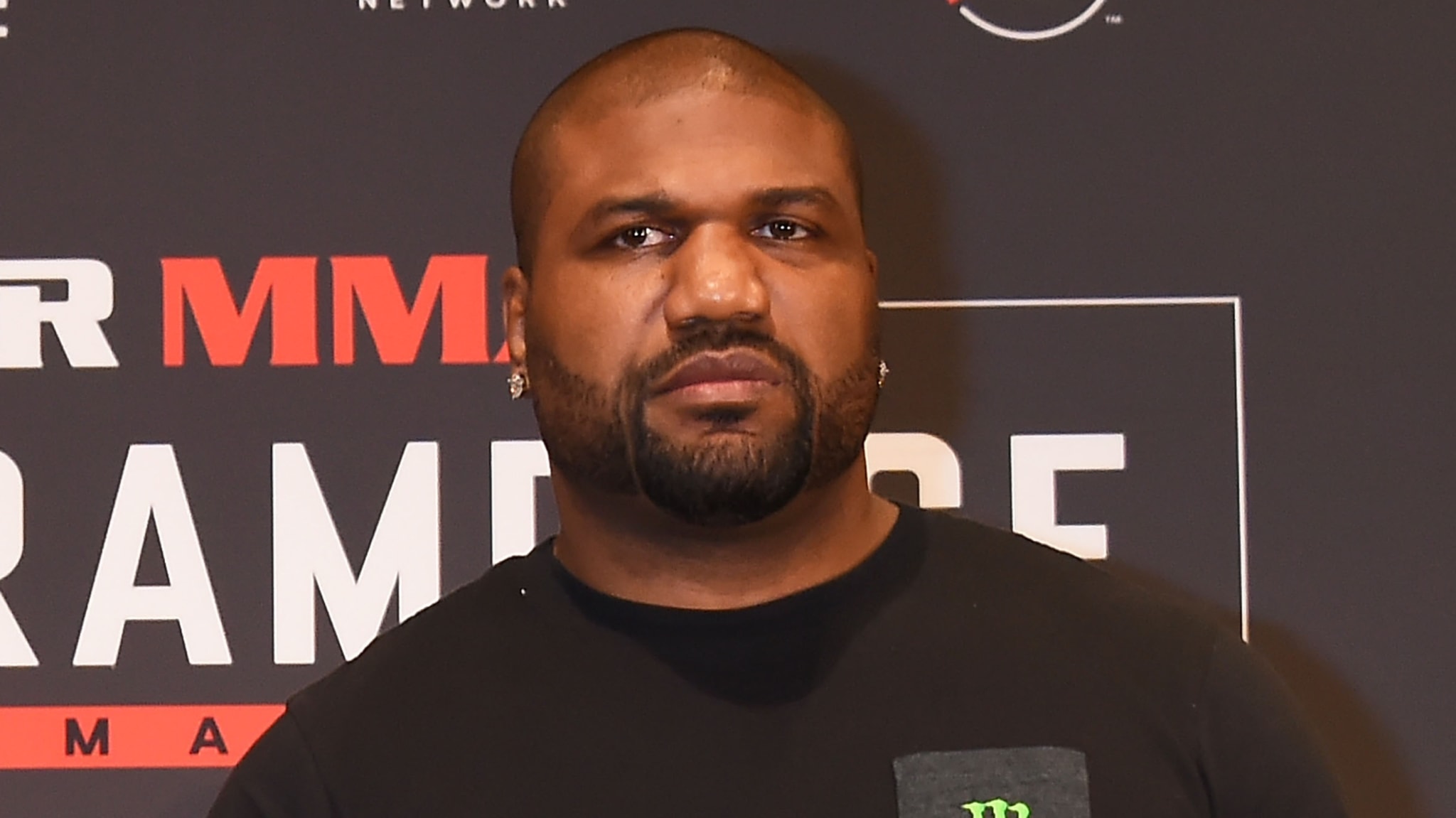A man with a serious expression stands in front of an MMA promotional backdrop.