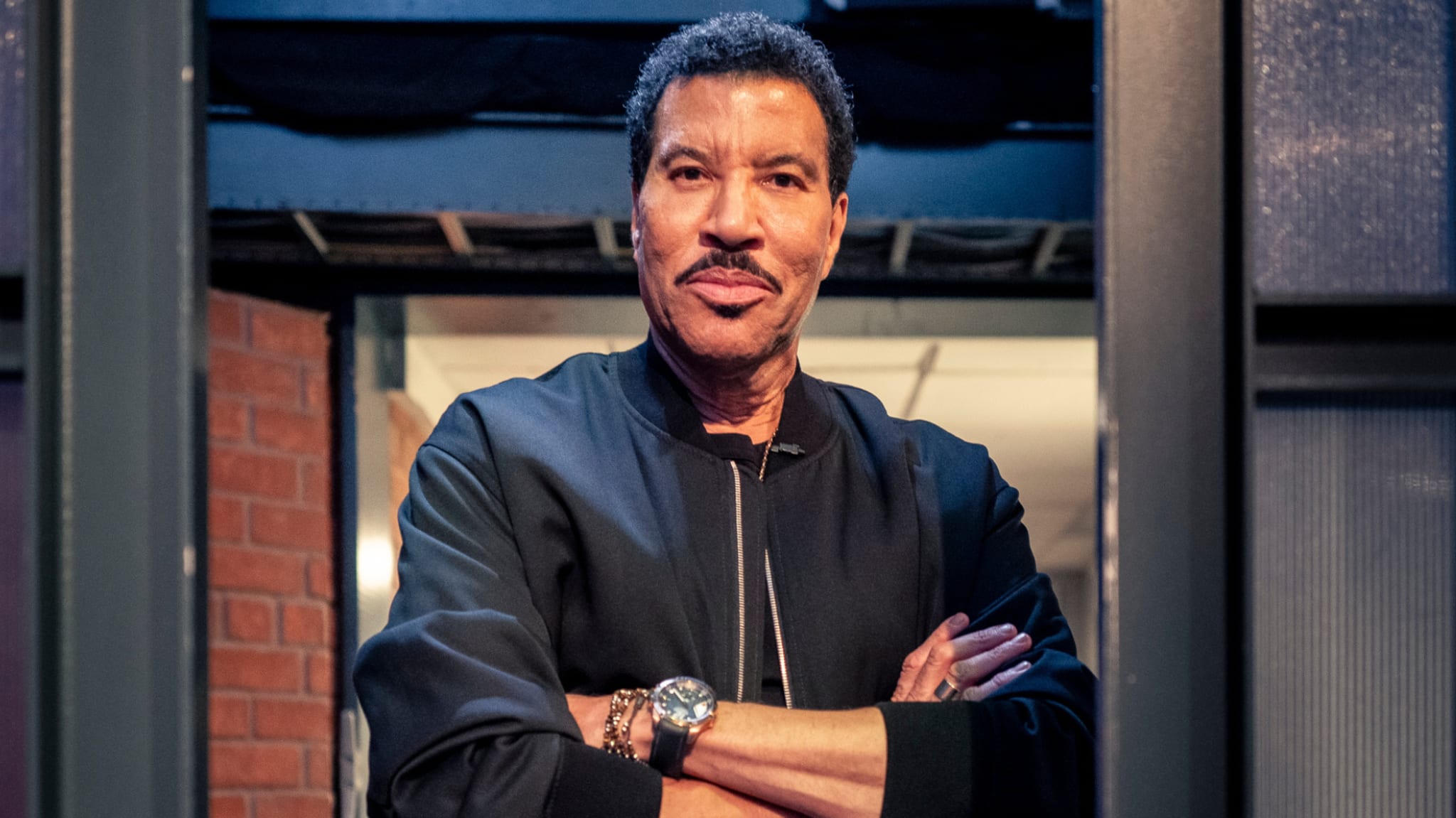 Lionel Richie, wearing a black jacket, stands with arms crossed in a doorway, smiling slightly.
