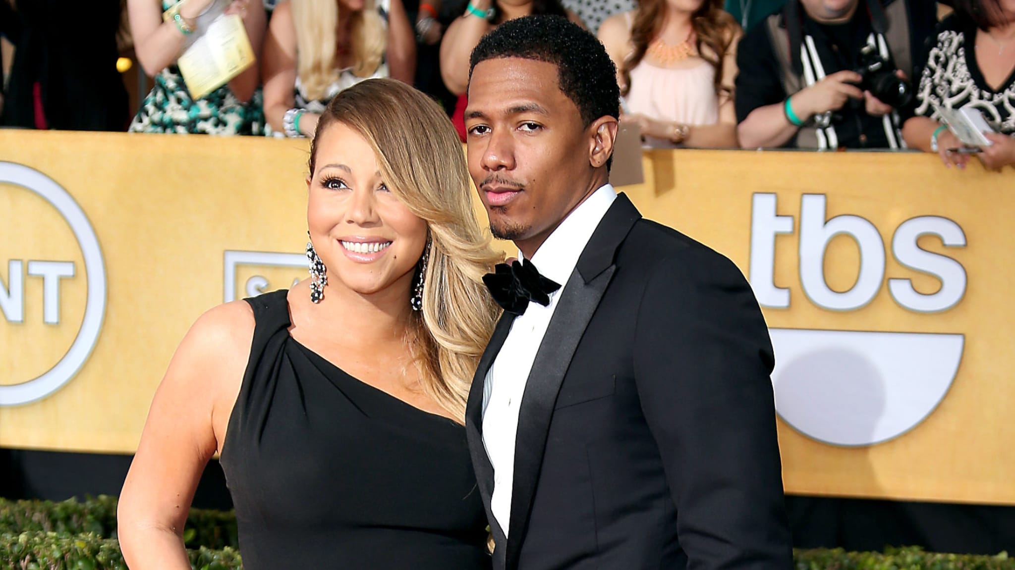 Mariah Carey and Nick Cannon pose together on a red carpet. Mariah wears a black dress, and Nick is in a tuxedo.