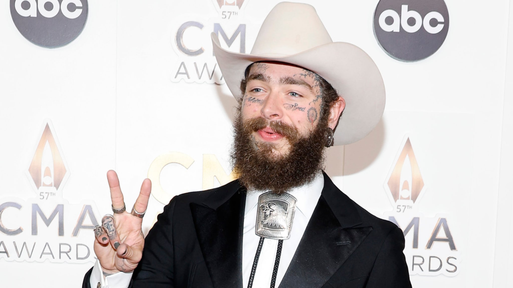 Post Malone at the 57th CMA Awards, wearing a cowboy hat and bolo tie, flashing a peace sign.