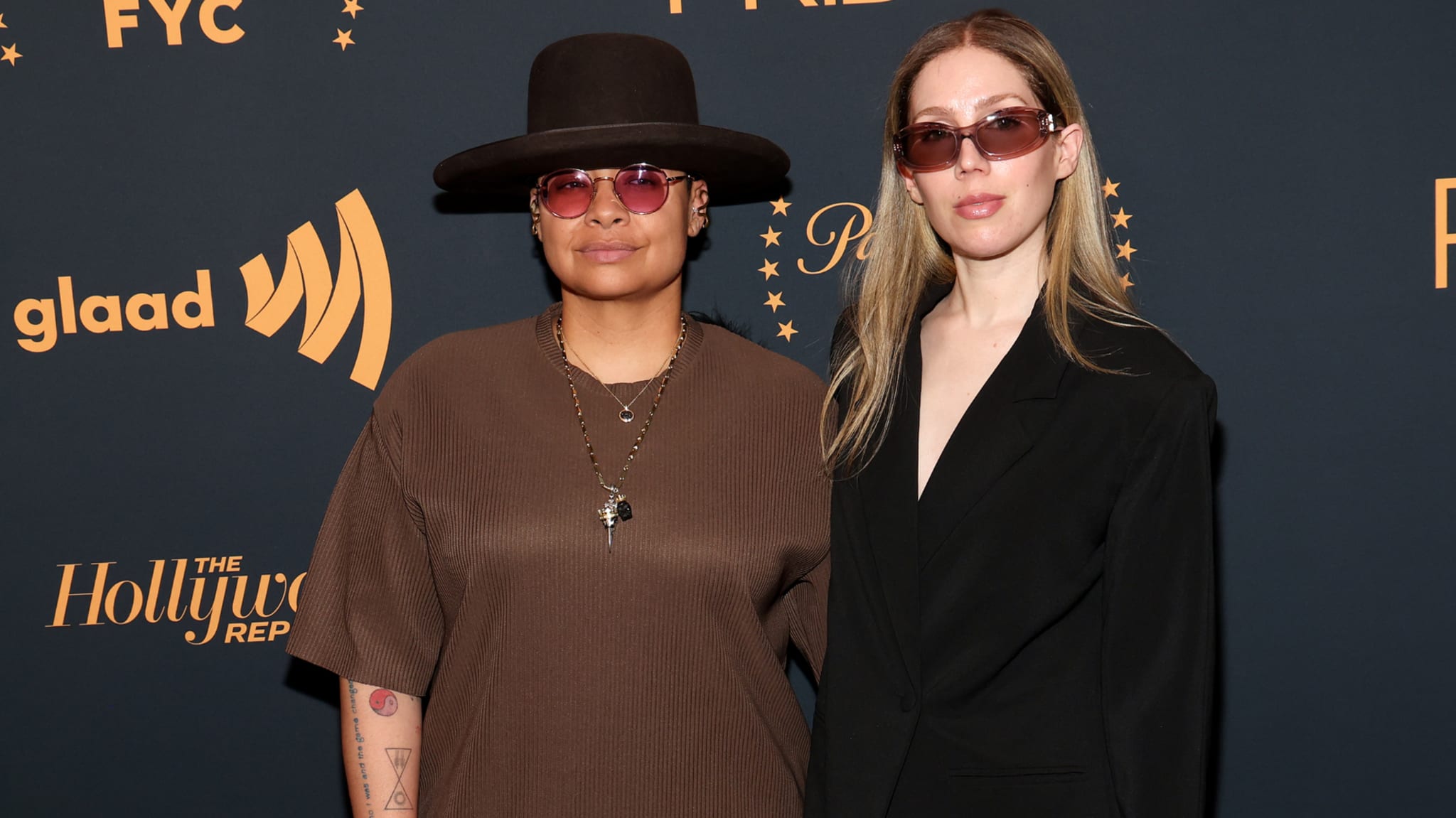 Two people posing on the red carpet at a GLAAD event, wearing sunglasses and stylish outfits.