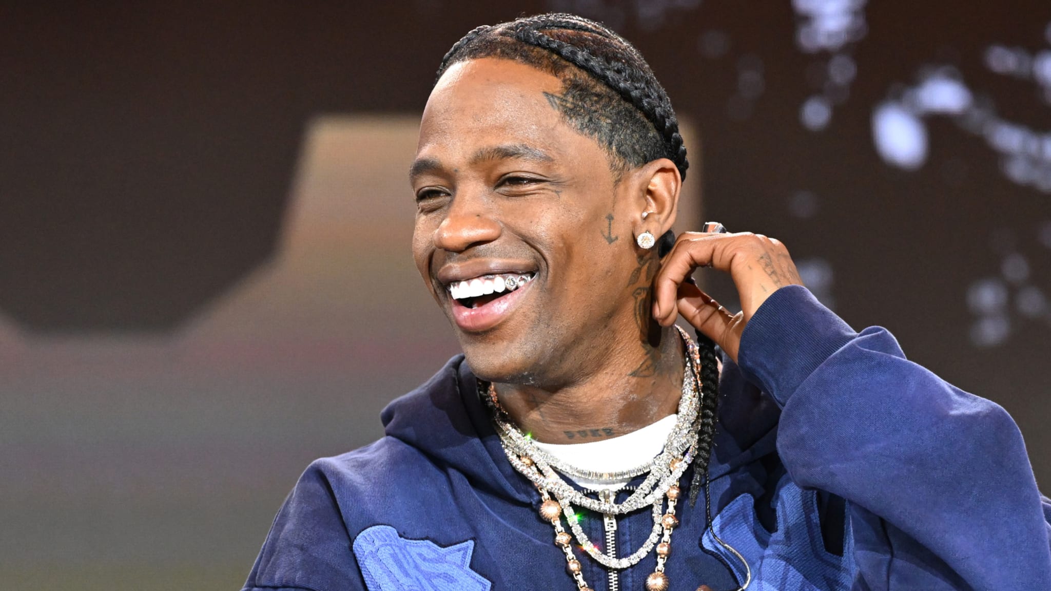 Travis Scott smiling, wearing a blue hoodie and multiple necklaces, with braided hair.