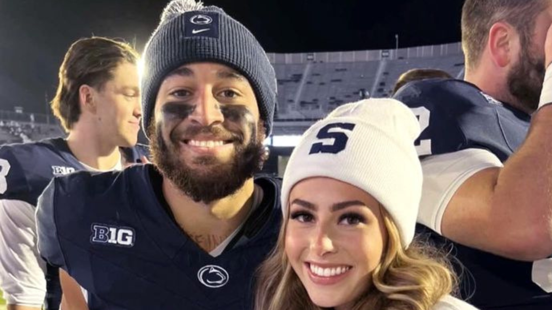 A man in a Penn State football uniform and a woman in a white beanie with an "S" are smiling at a stadium.