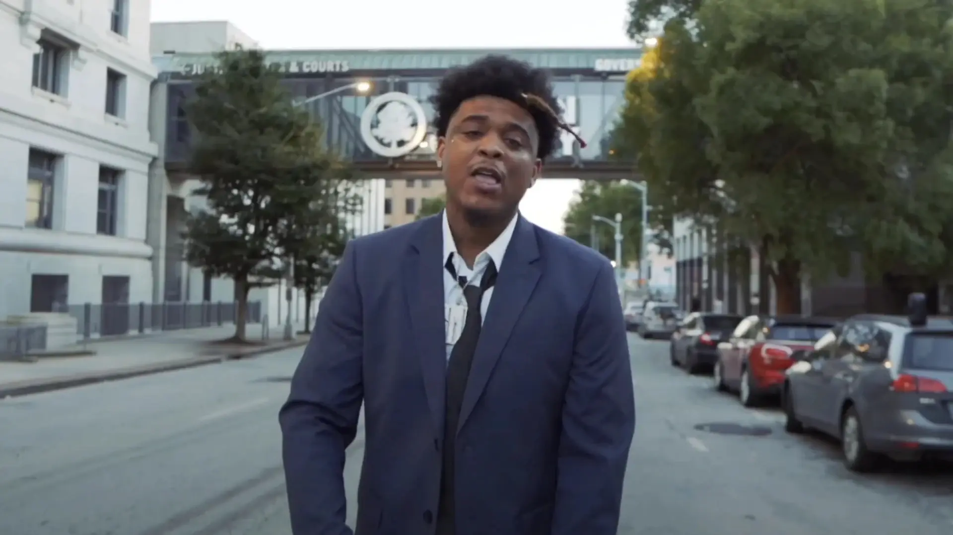 A man in a suit stands on an empty city street with parked cars. A building and a bridge are in the background.