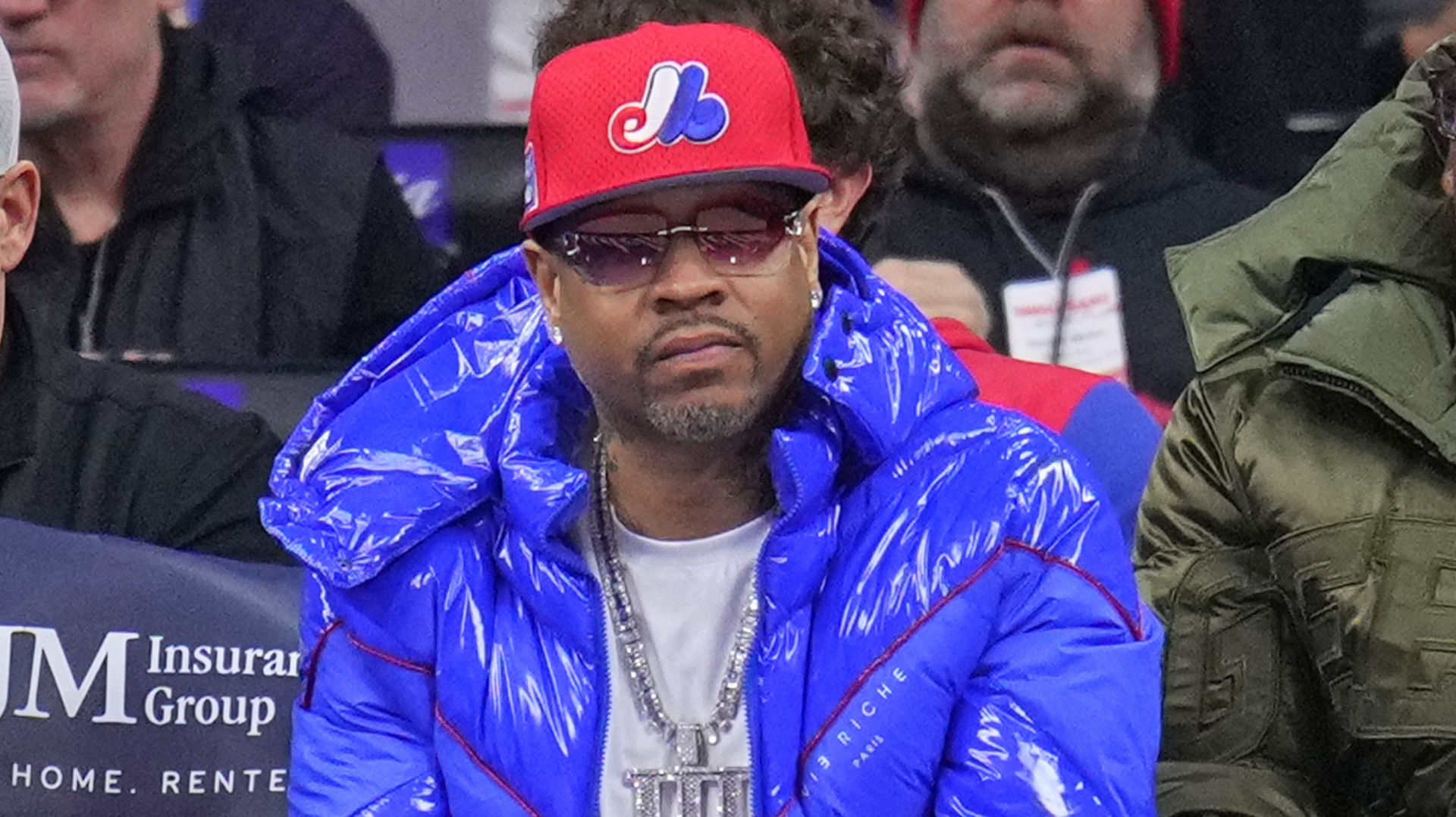 A person wearing a red baseball cap, sunglasses, and a blue puffer jacket sits in a crowd.