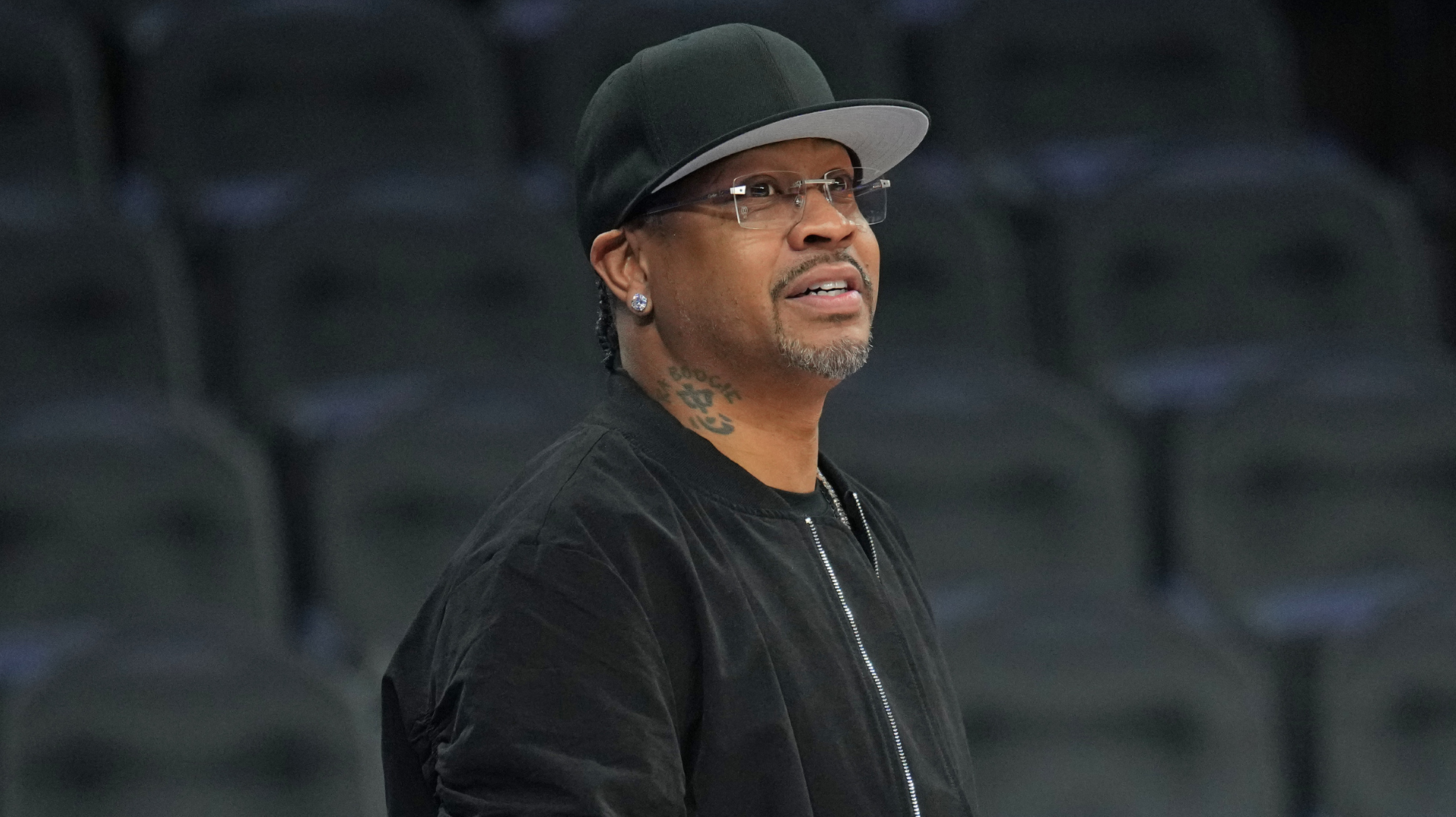A man wearing a black cap, glasses, and a black jacket is standing in an empty stadium.