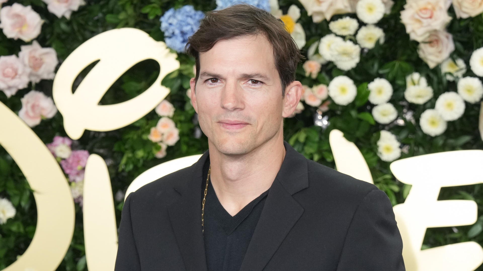 A man in a black suit stands in front of a floral background with a large "D" logo.