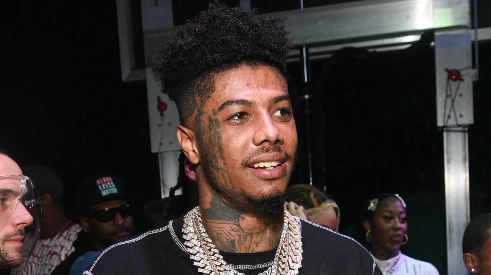 Rapper Blueface is seen wearing a black shirt with silver chains, smiling at an event.