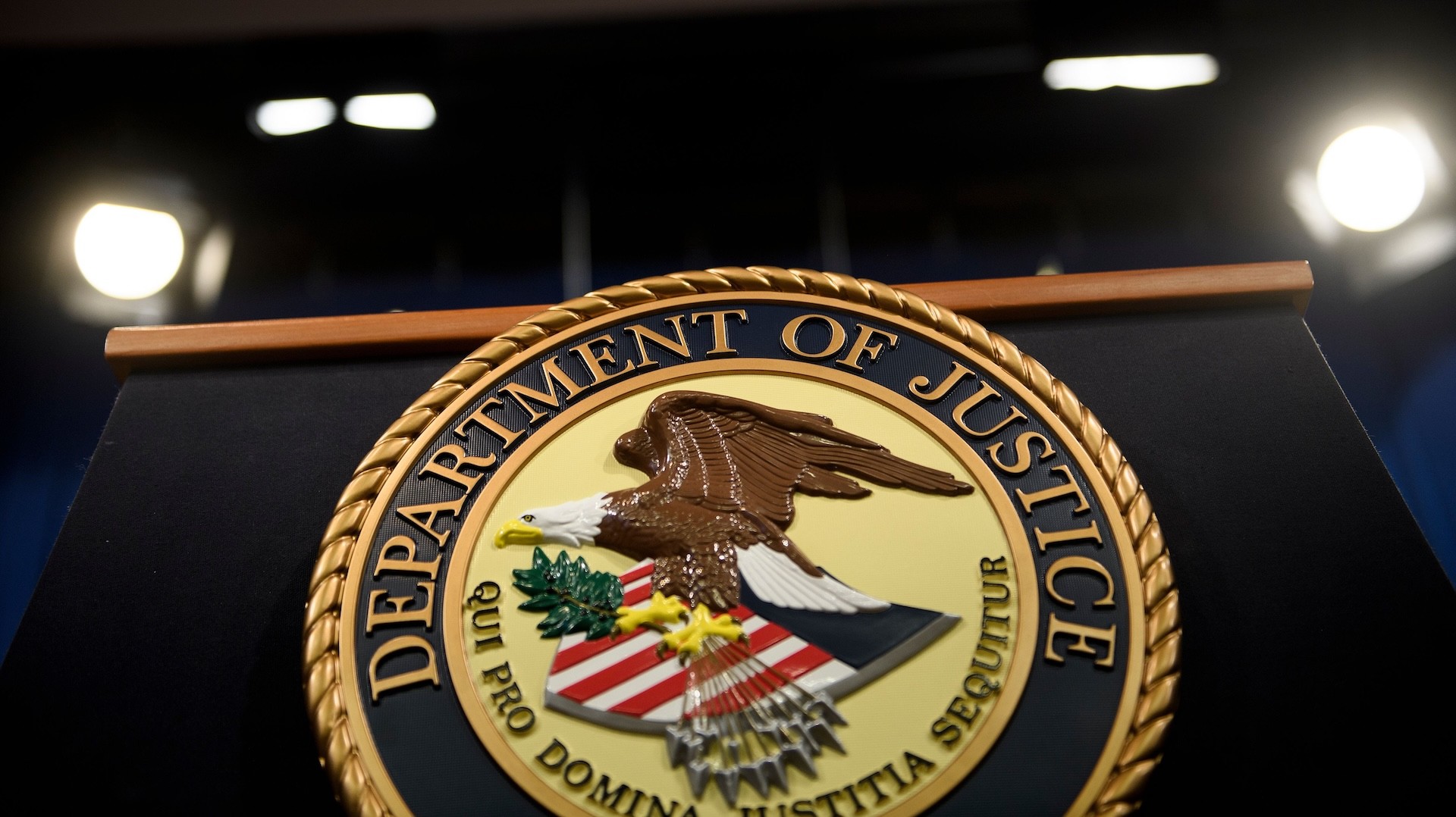 Seal of the United States Department of Justice on a podium, featuring an eagle and shield, with bright lights in the background.