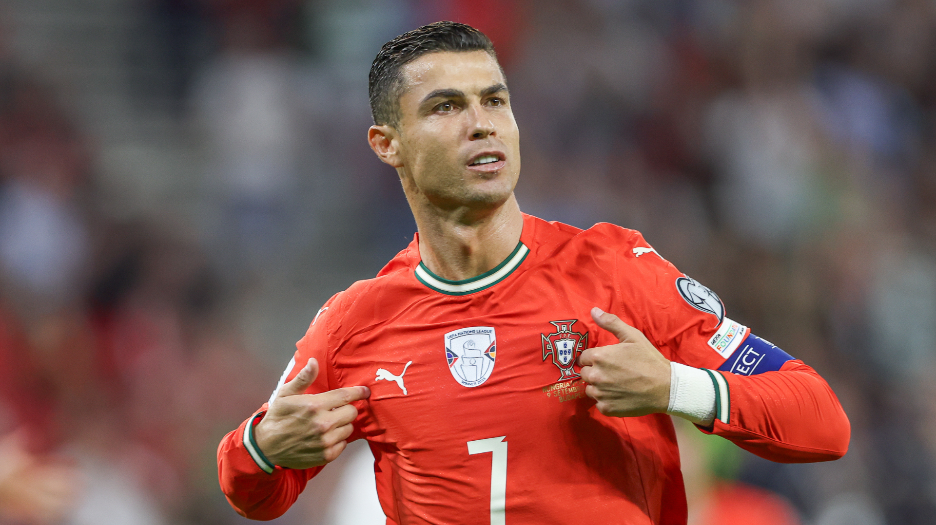 Cristiano Ronaldo in a red Portugal jersey, pointing to himself, wearing a captain's armband, on a soccer field.