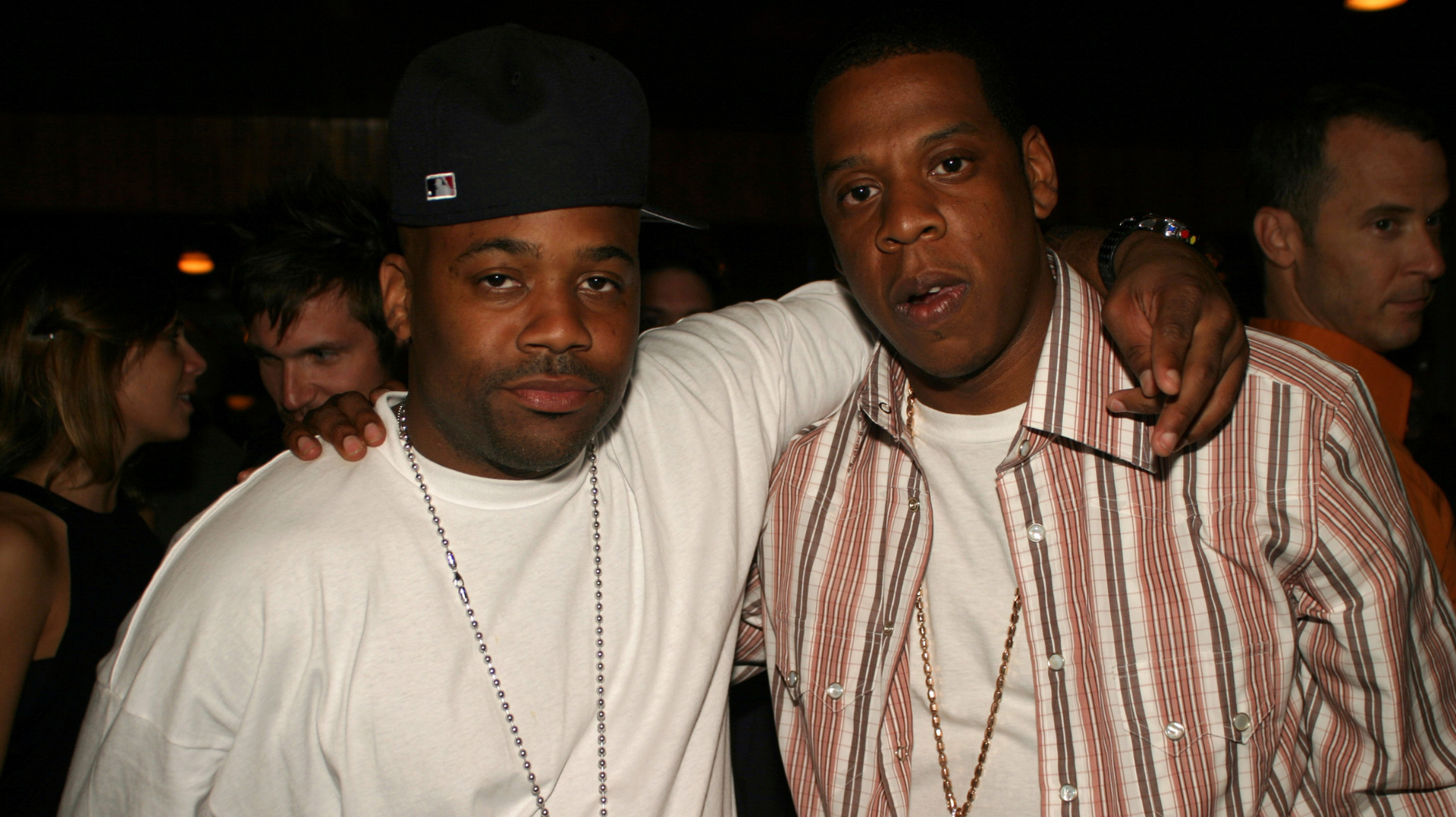 Damon Dash and Jay-Z pose together at an event. Damon wears a white shirt and cap, while Jay-Z sports a striped shirt and chain.