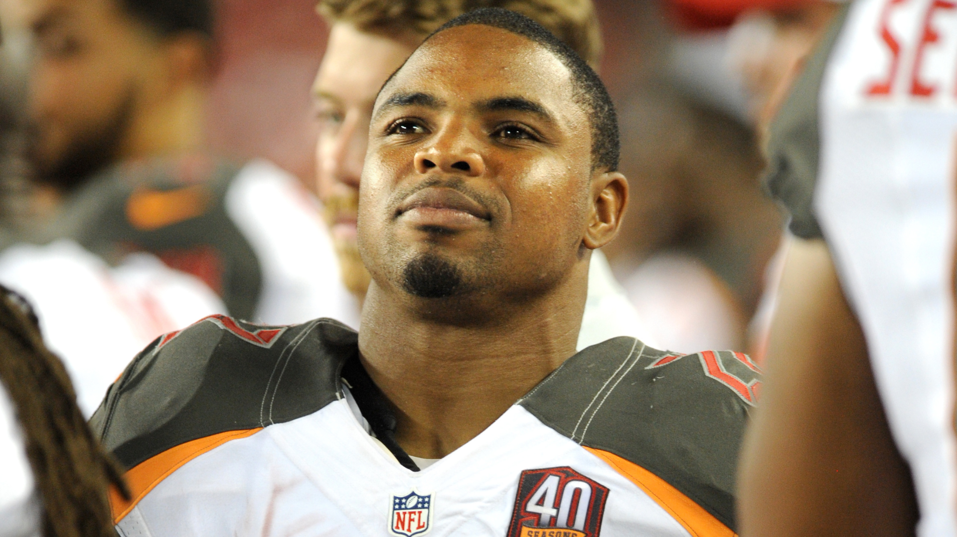 A football player in a Tampa Bay Buccaneers uniform on the sidelines, looking thoughtful.