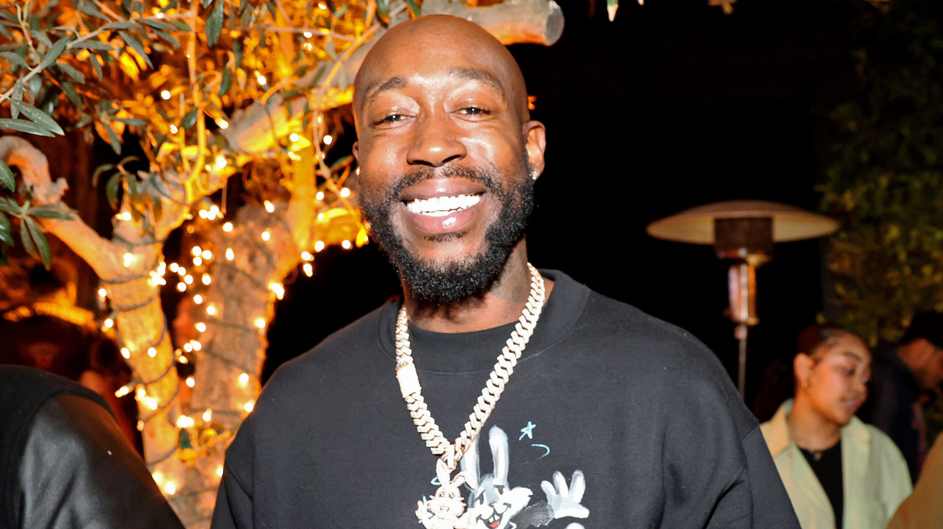 Freddie Gibbs smiling at an event, wearing a black shirt and a gold chain, with decorative lights in the background.