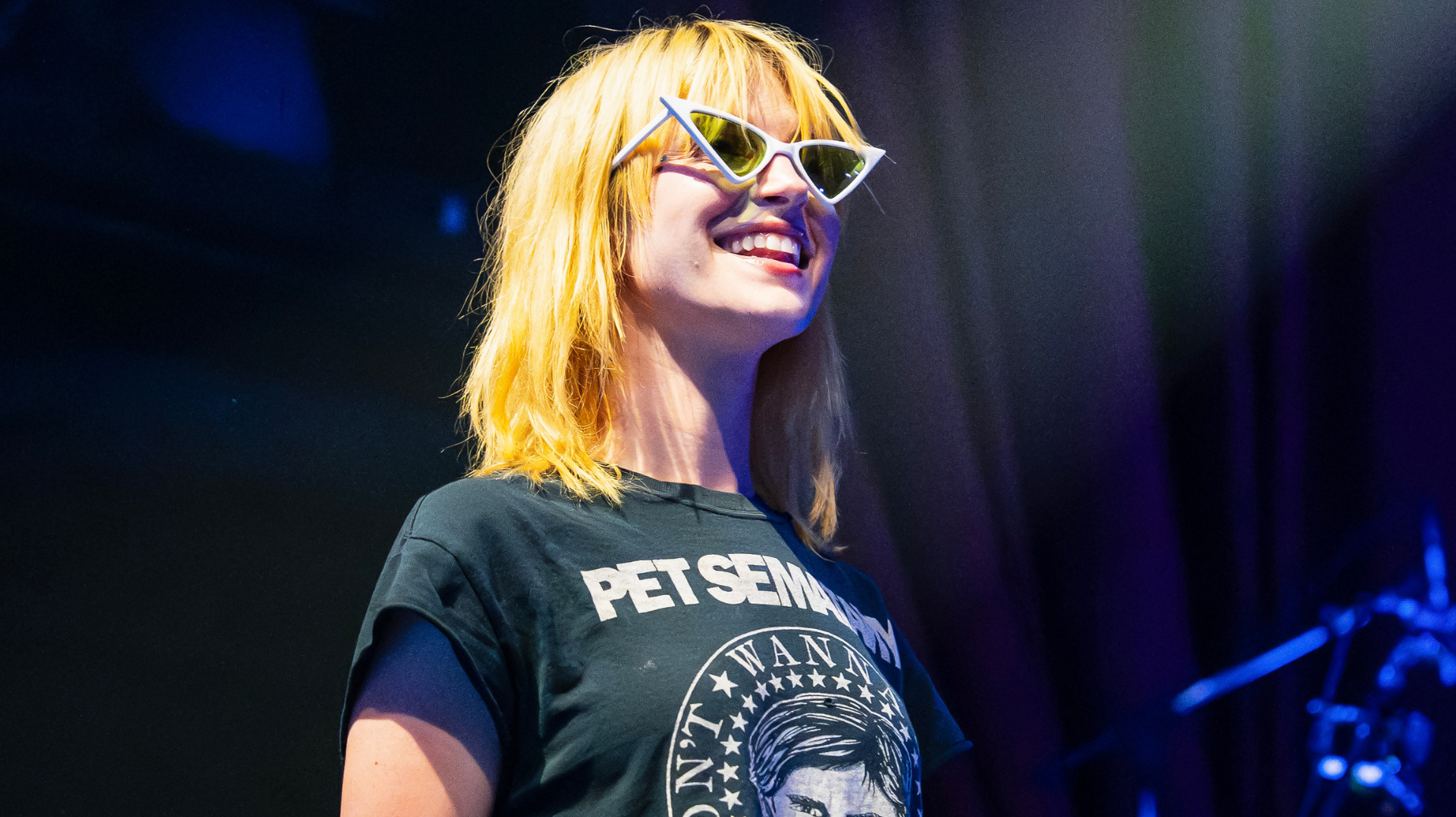 A person with blonde hair wearing cat-eye sunglasses and a "Pet Sematary" shirt, smiling on stage.
