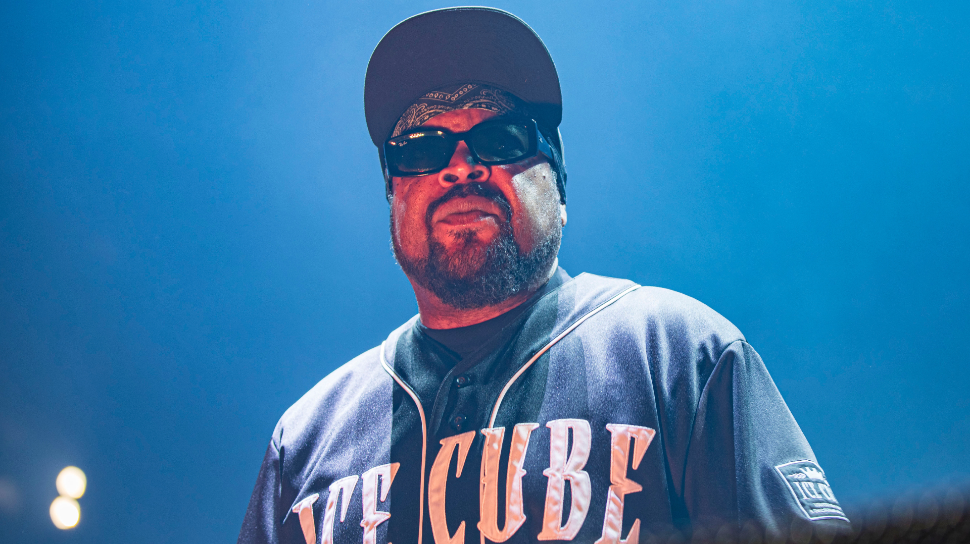 Ice Cube on stage wearing sunglasses and a cap, with a serious expression, under blue lighting.
