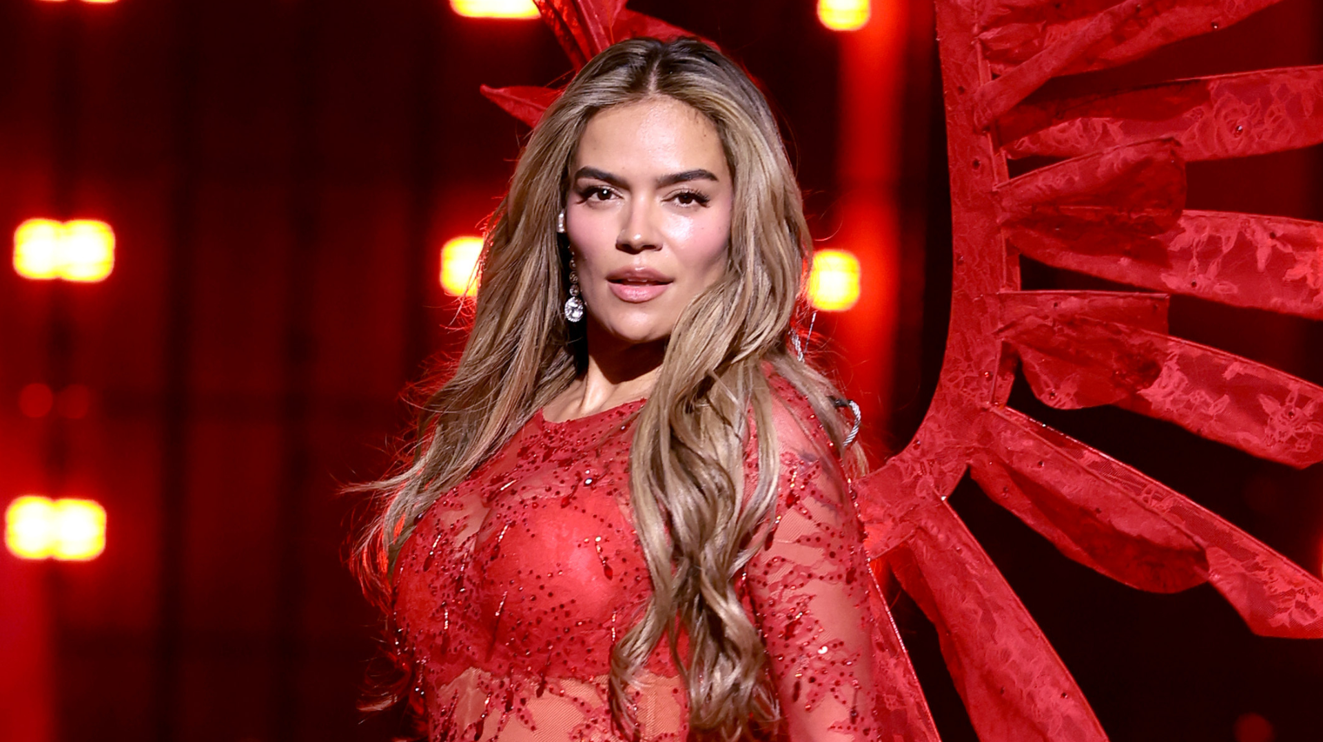 A woman in a red, sparkling outfit with long, wavy hair stands confidently on stage with red lights in the background.