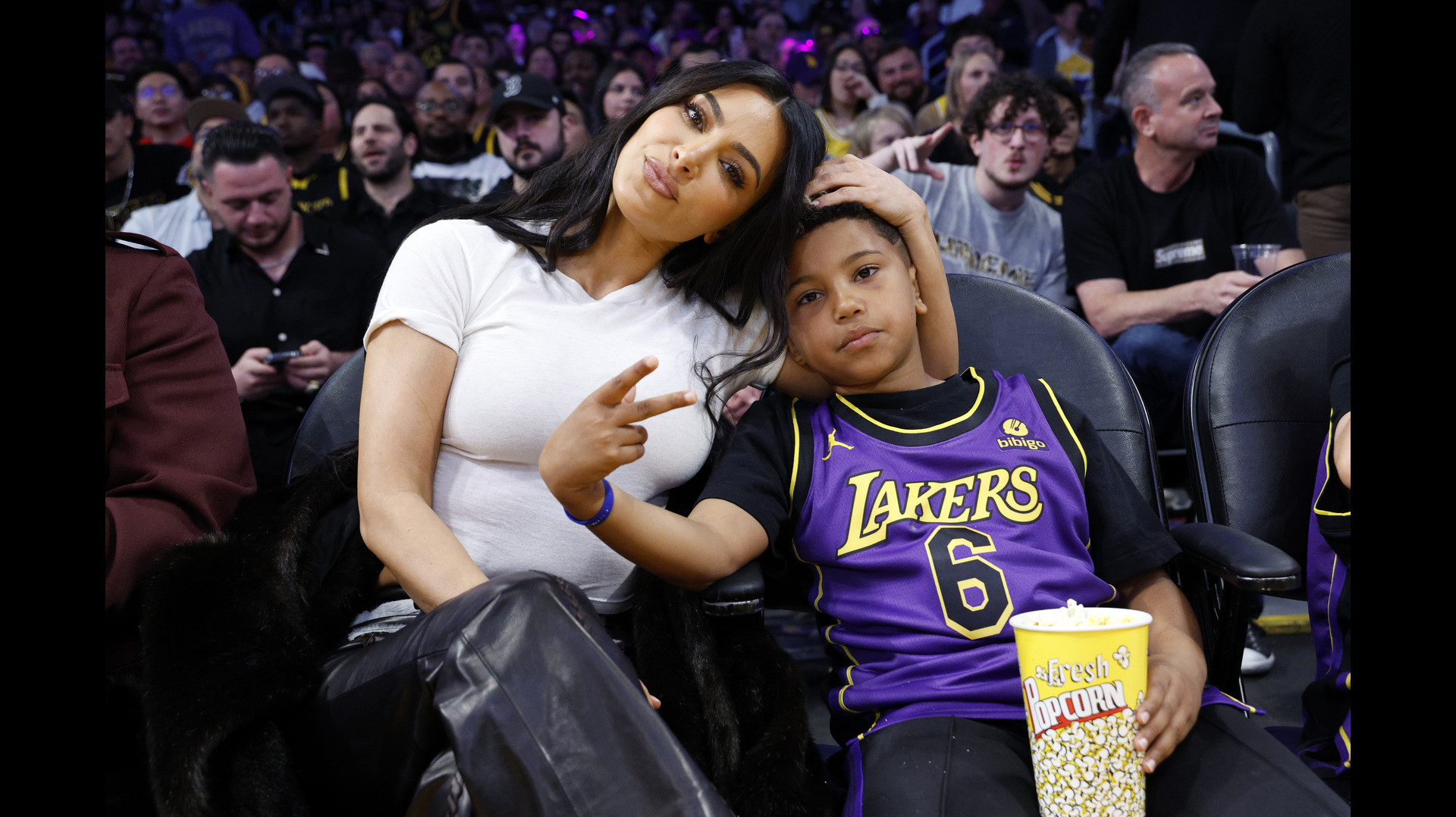 Kim Kardashian sitting courtside with her son at a Lakers game, both posing for the camera. Saint is making a peace sign.