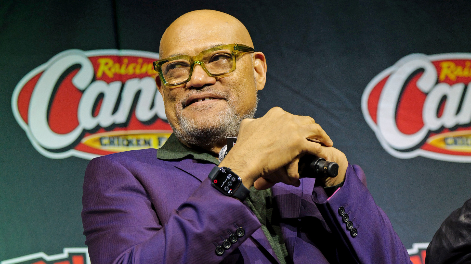 Laurence Fishburne in a purple suit, wearing glasses, smiles while holding a microphone at an event with a Raising Cane's backdrop.