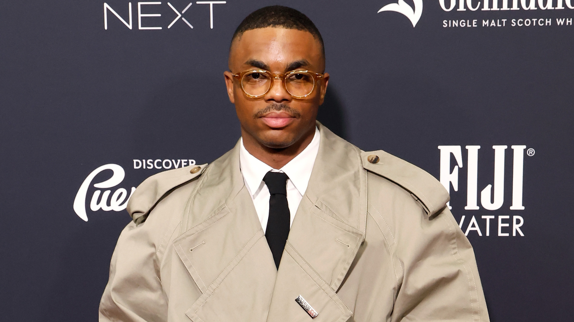 A man in glasses and a beige trench coat stands in front of a branded backdrop.
