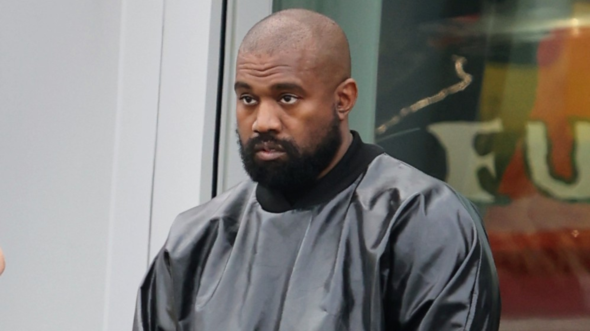 Kanye West in a black outfit, standing in front of a window with a serious expression.