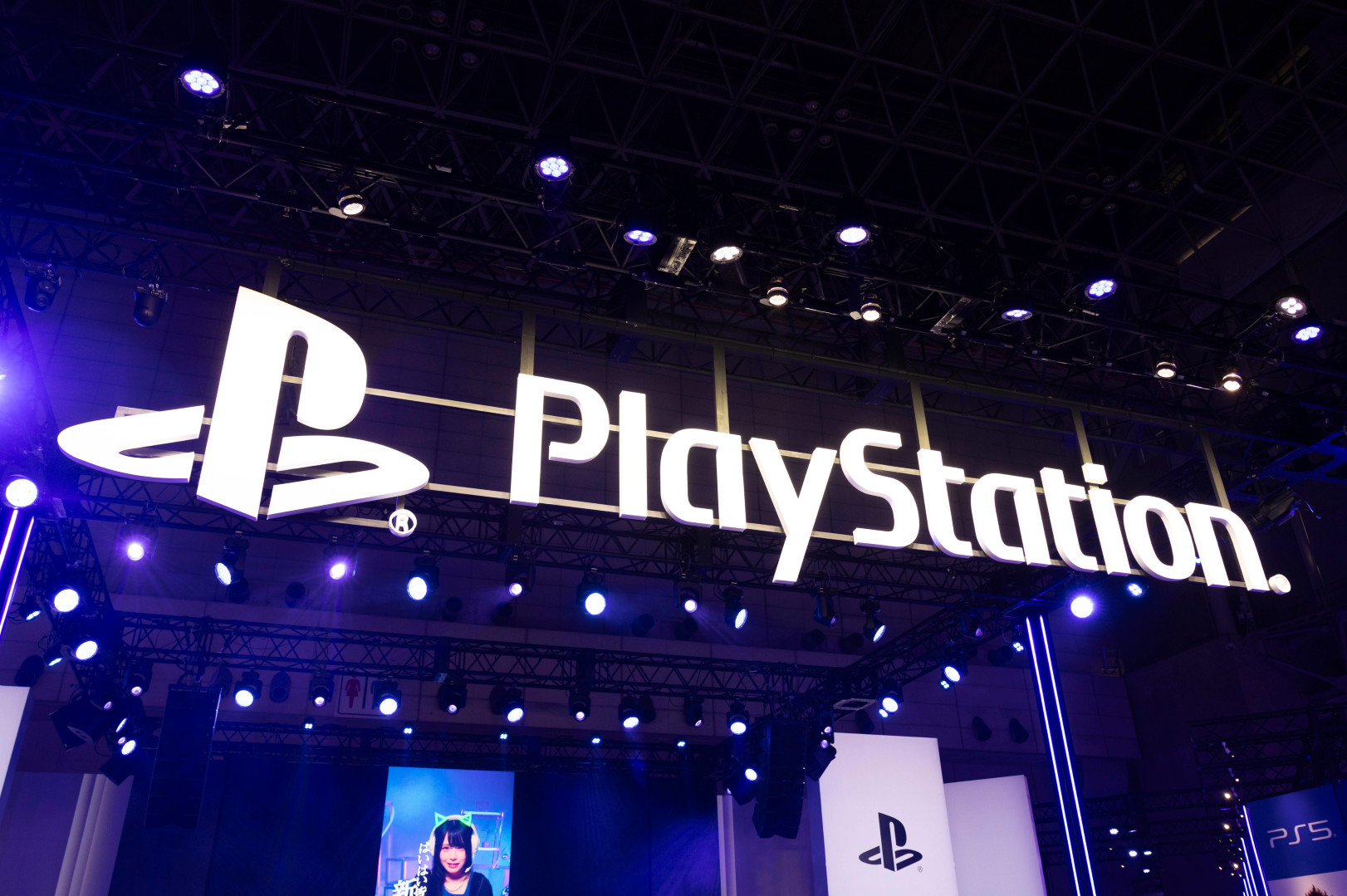 PlayStation booth with a large illuminated logo at a gaming event, featuring a stage and screens displaying content.
