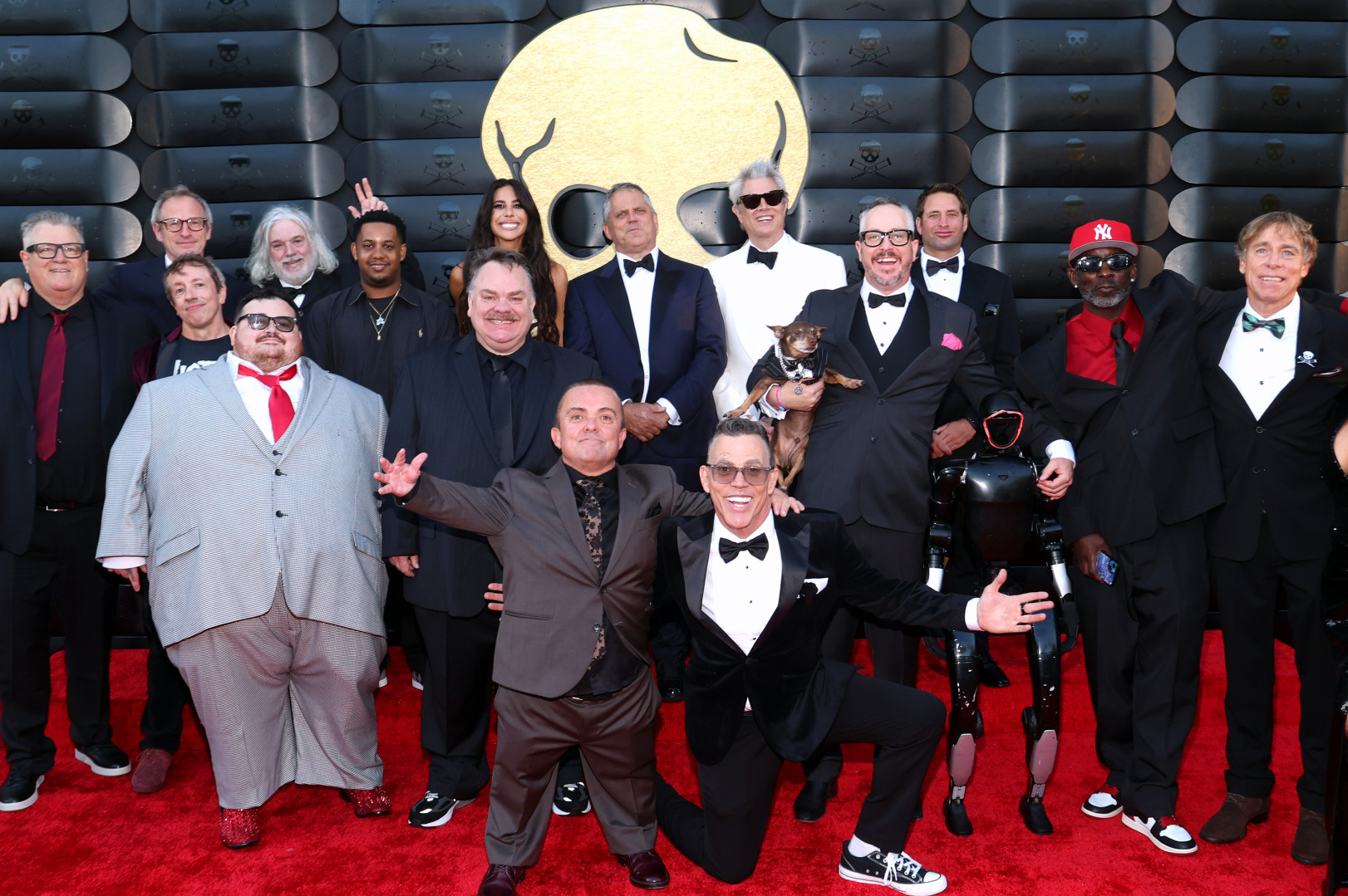 A group of stars, including the cast of "Jackass," pose on a red carpet in formal attire with a large logo in the background.
